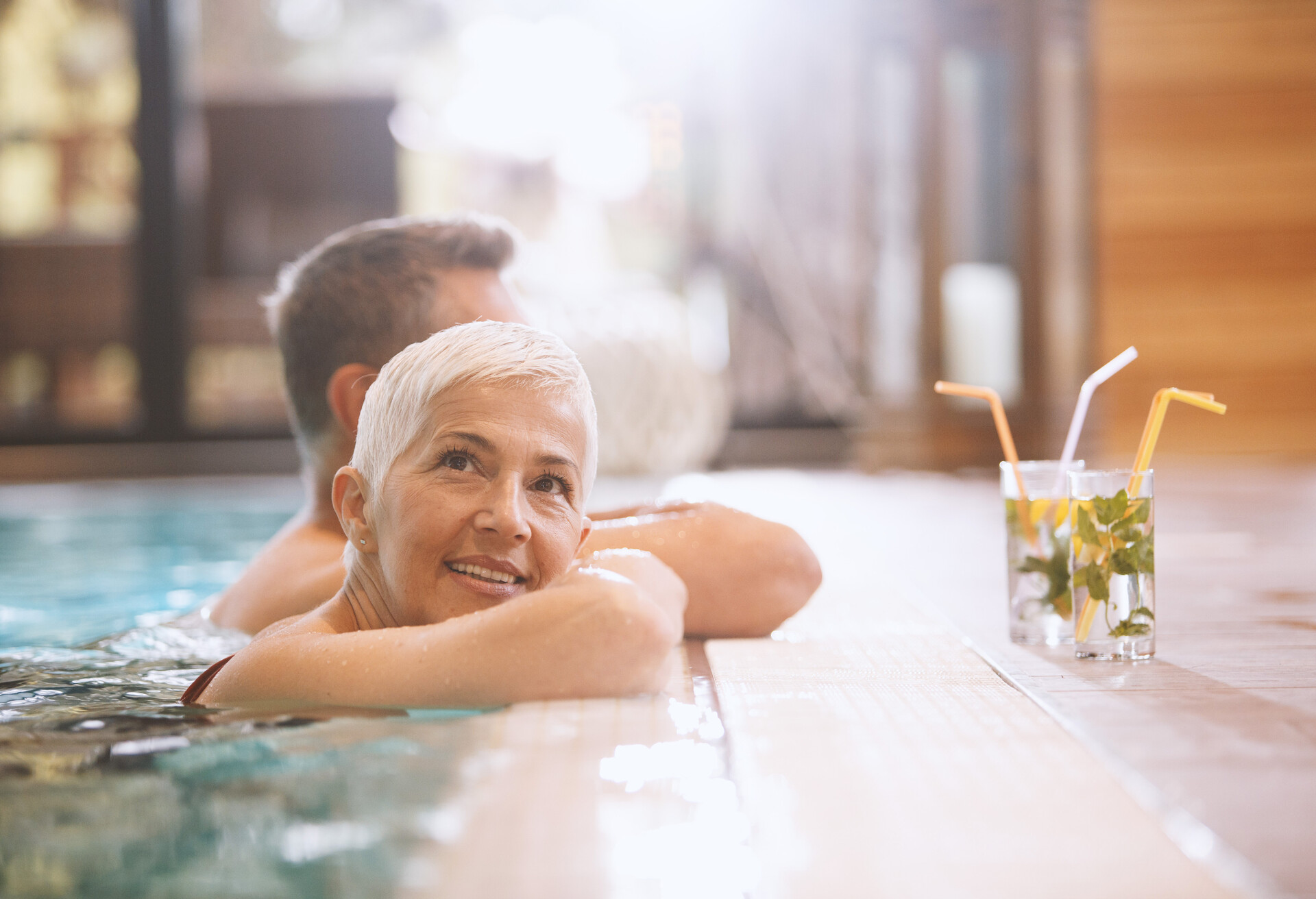 Senior woman and her husband are enjoying the day at the spa. They are relaxing in the indoor swimming pool, having cocktails and enjoying the sun shining through windows.