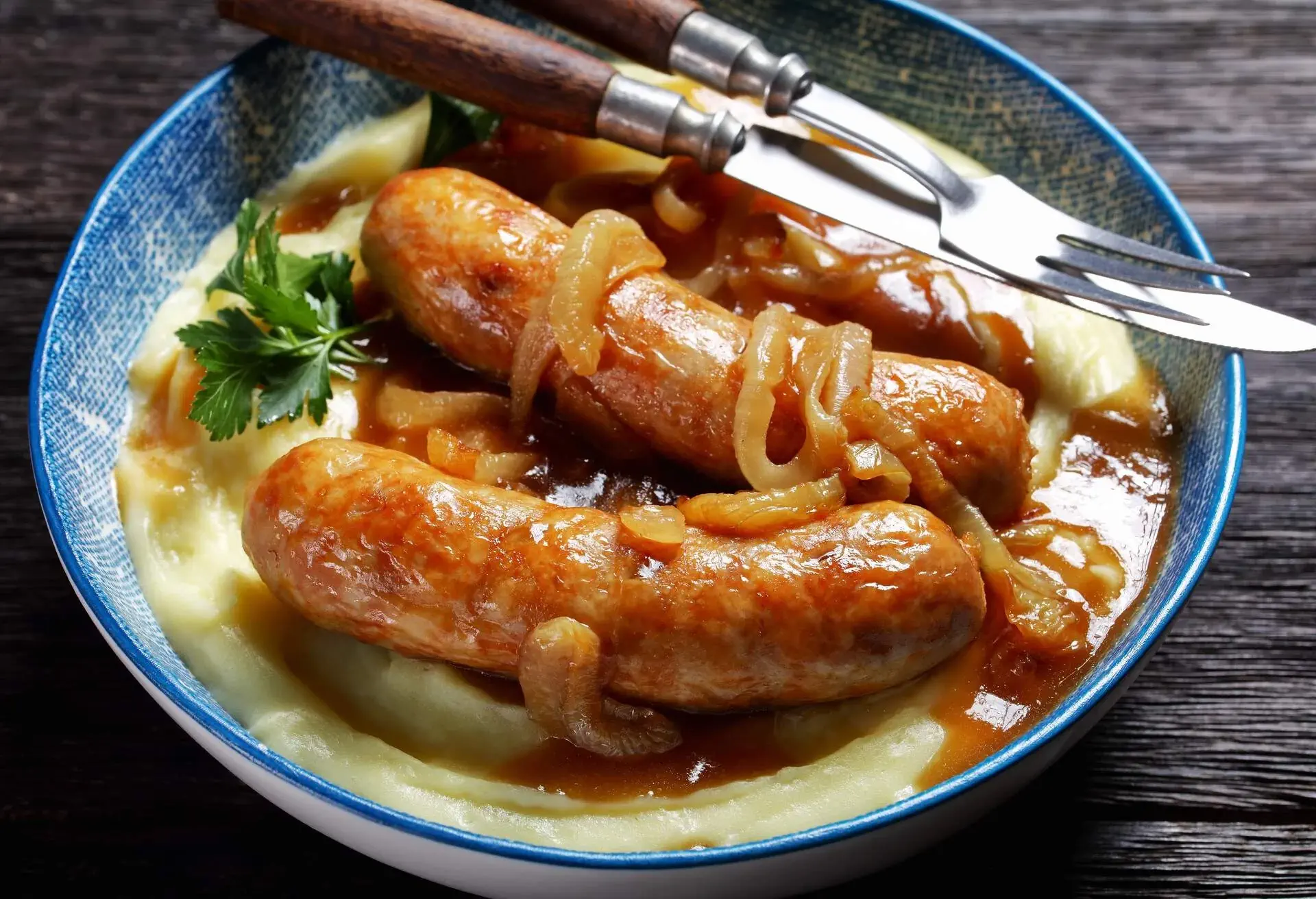 Bangers and mash traditional british dish of pork sausage with onion gravy and mashed potato on a blue plate with cutlery on a dark wooden background, top view, close-up