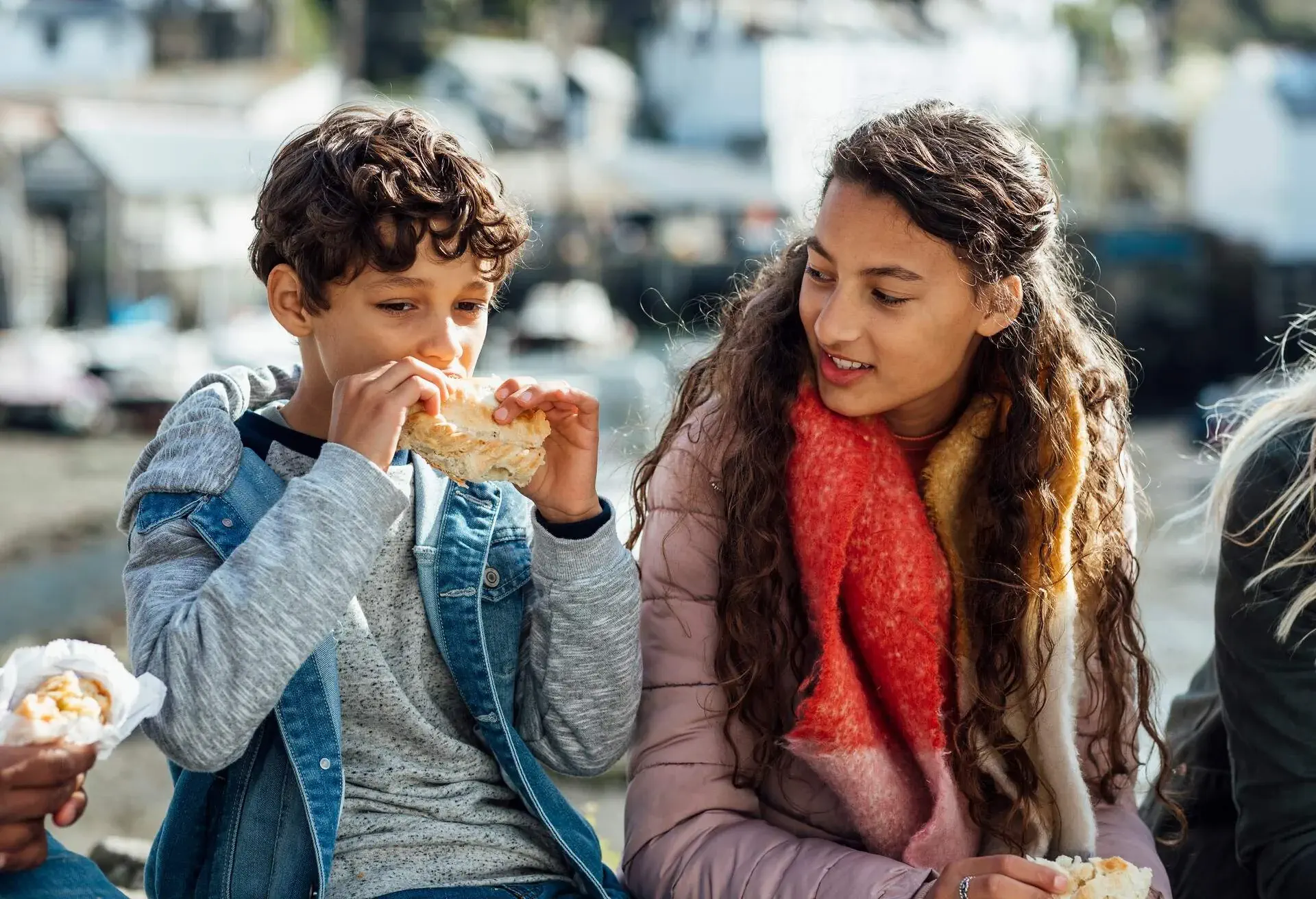 Mixed race brother and sister sitting on a stone wall in Polperro, Cornwall. They are eating Cornish pasties from local businesses while talking with each other. 