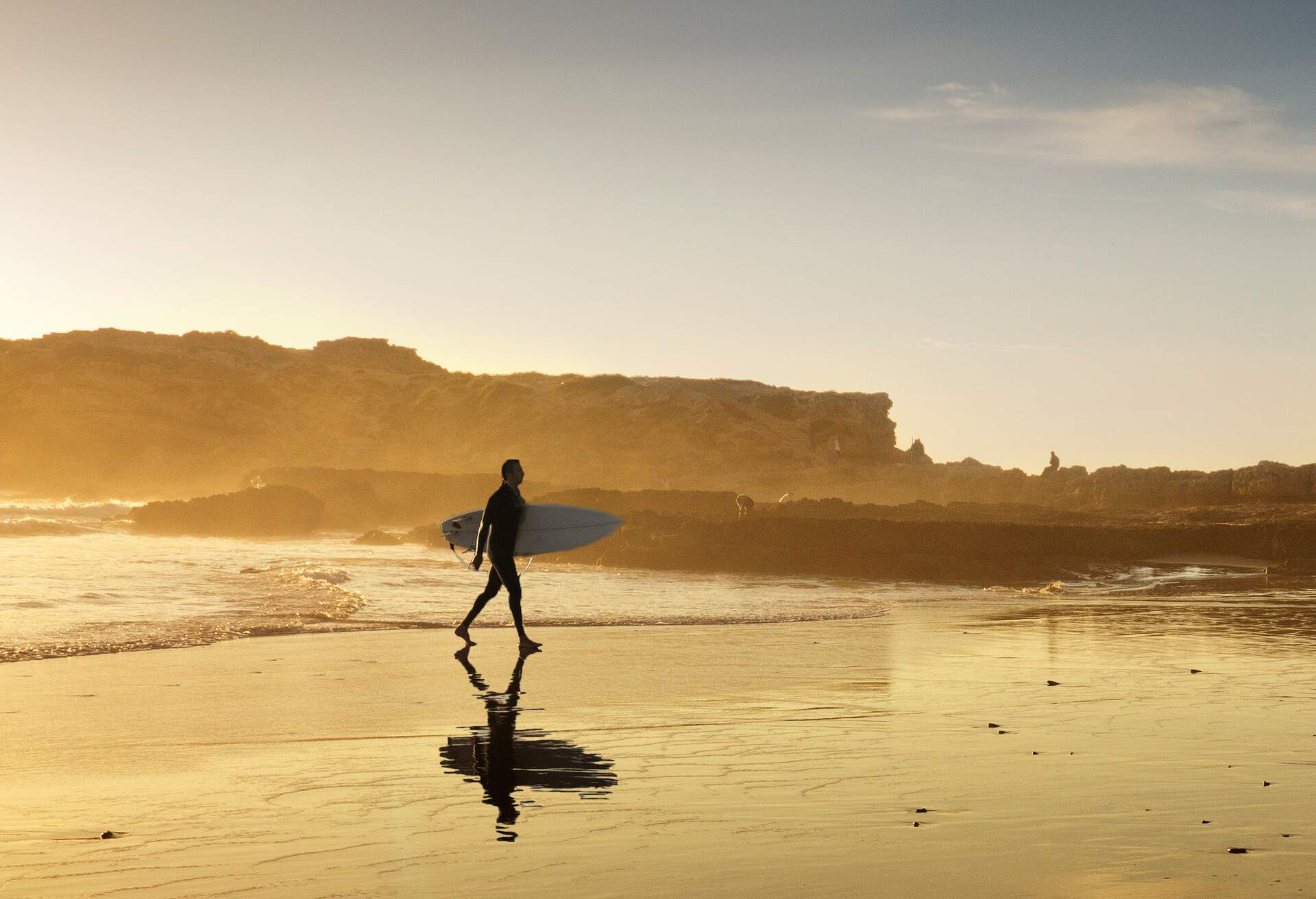 A surfer in golden sunset light is coming out of the water. His silhouette is reflecting on the beach
