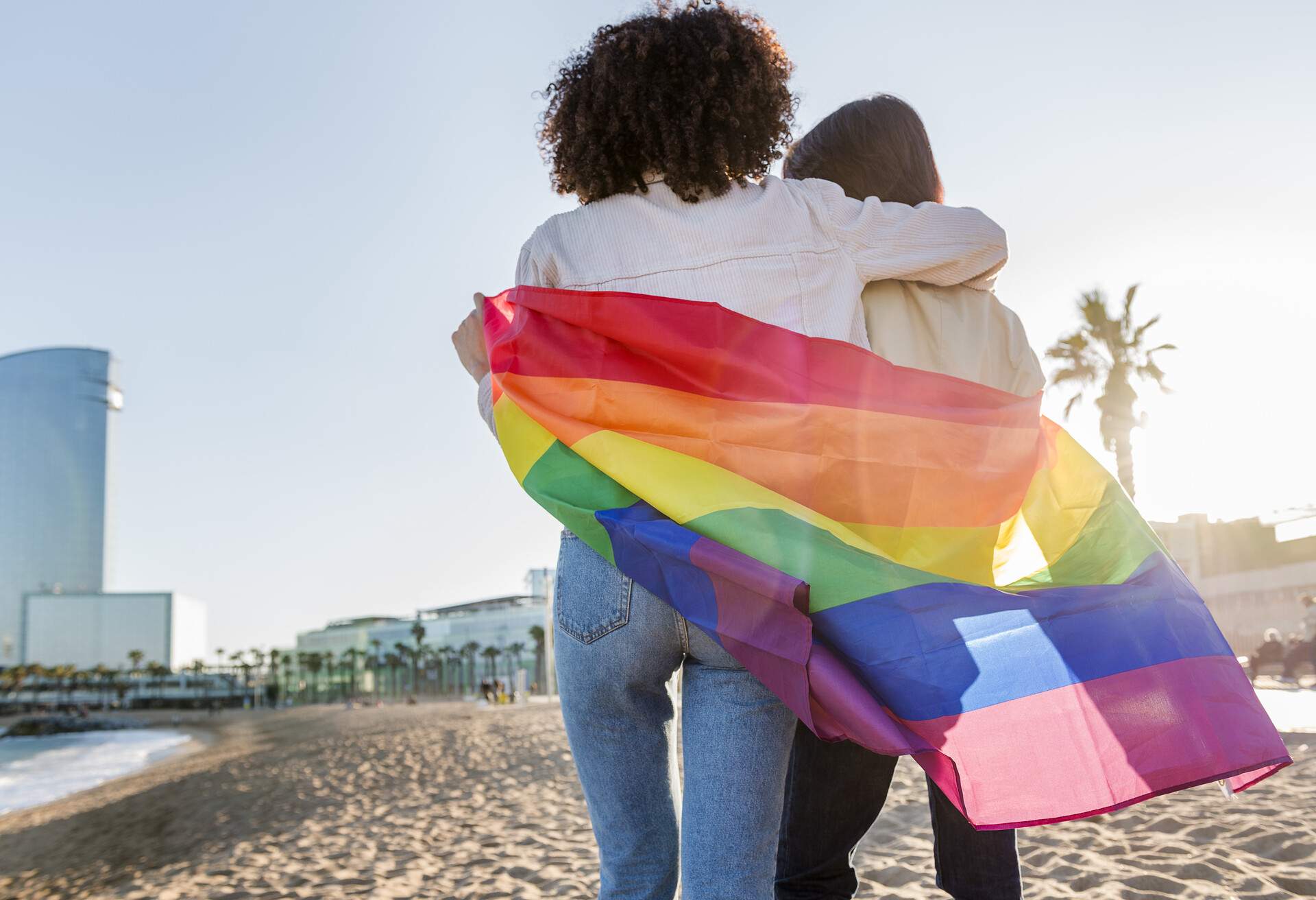 interracial couple of gay girls walking in front of the sea at Barcelona wrapped in the rainbow flag, symbol of the struggle for homosexual rights, concept of sexual freedom and racial diversity