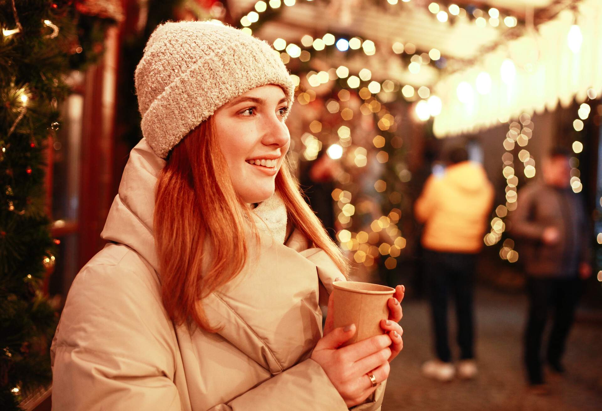 Happy young woman at Christmas market in the winter time. Christmas lights and tree decorations on street. Holidays concept.