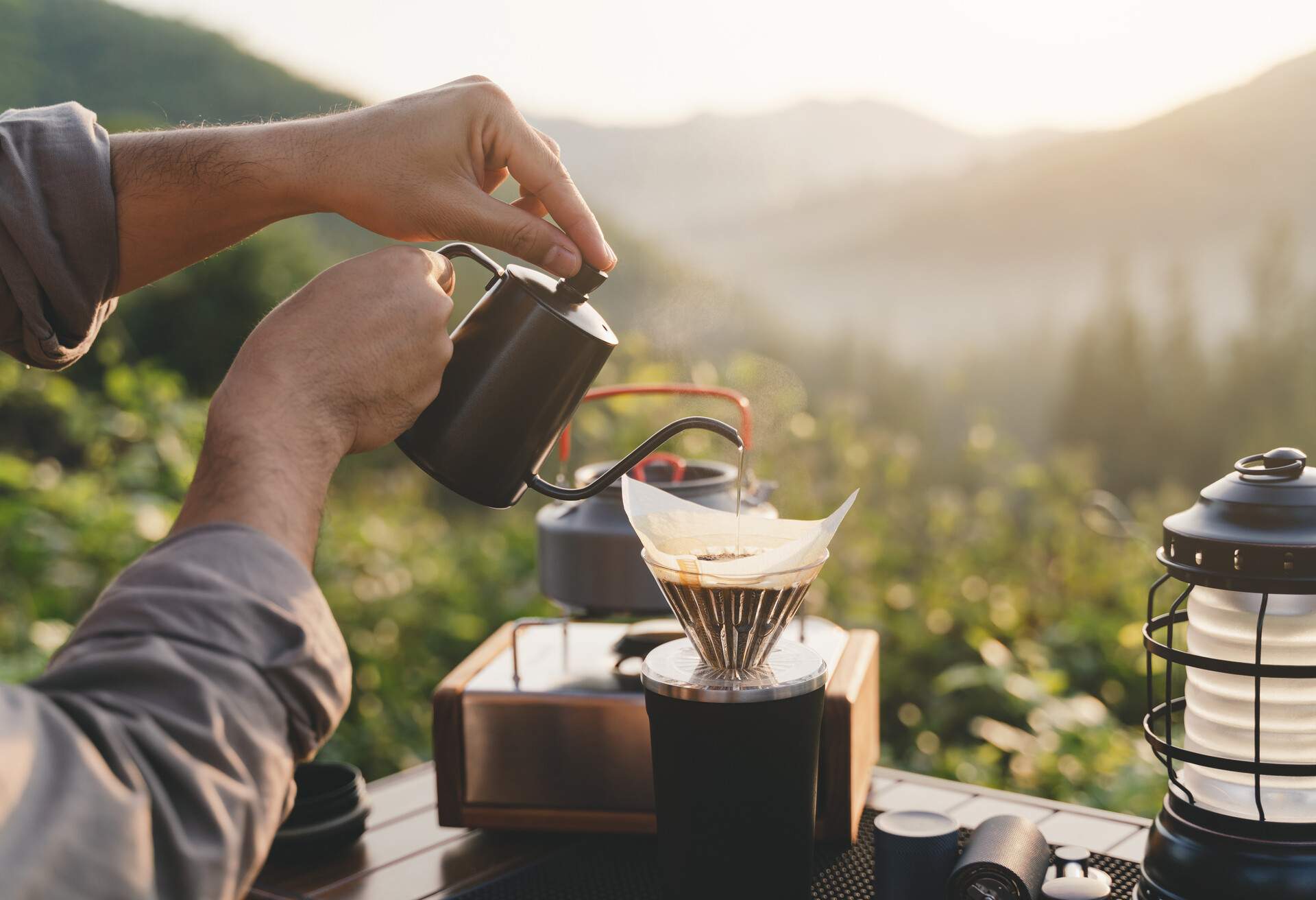 Rearview of a man making dripping coffee on the picnic table while camping on th mountain in morning