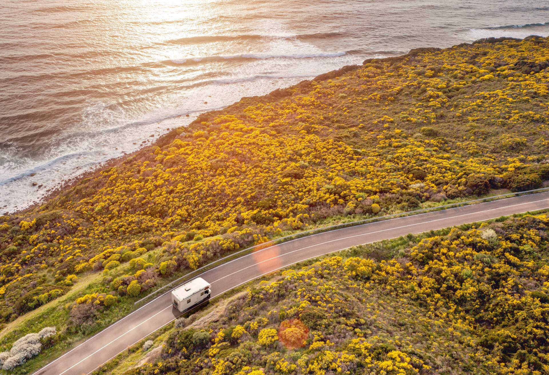 Driving a motor home on a road by the sea at sunset, with warm atmosphere and lens flare, seen from the air. Beautiful coastal scenery with sea waves breaking on a beach. Camping life, alternative lifestyle, freedom.