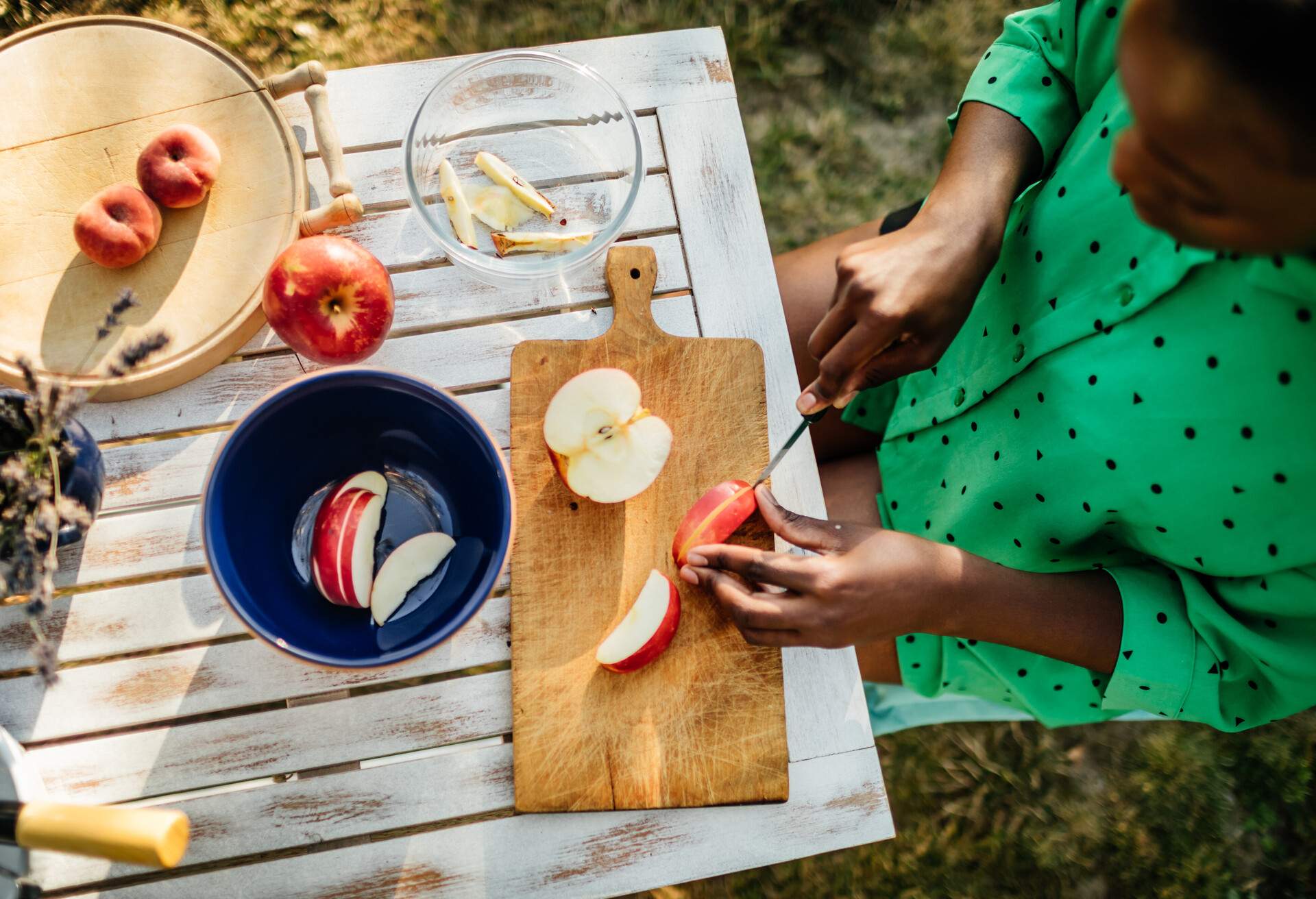 A woman cutting fruit at a wooden table outdoors preparing a snack for her family in the afternoon sun.