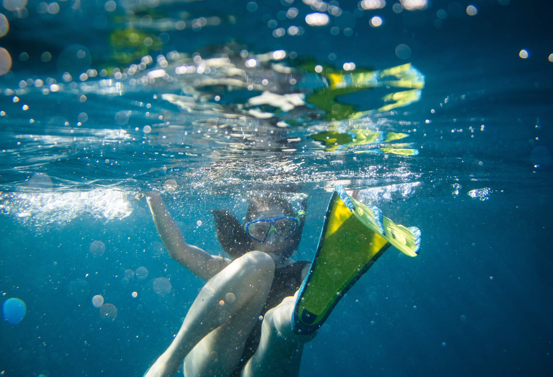 Woman with swimming goggles and diving fins diving looking at camera while diving underwater in the sea surrounded by air bubbles, water surface above