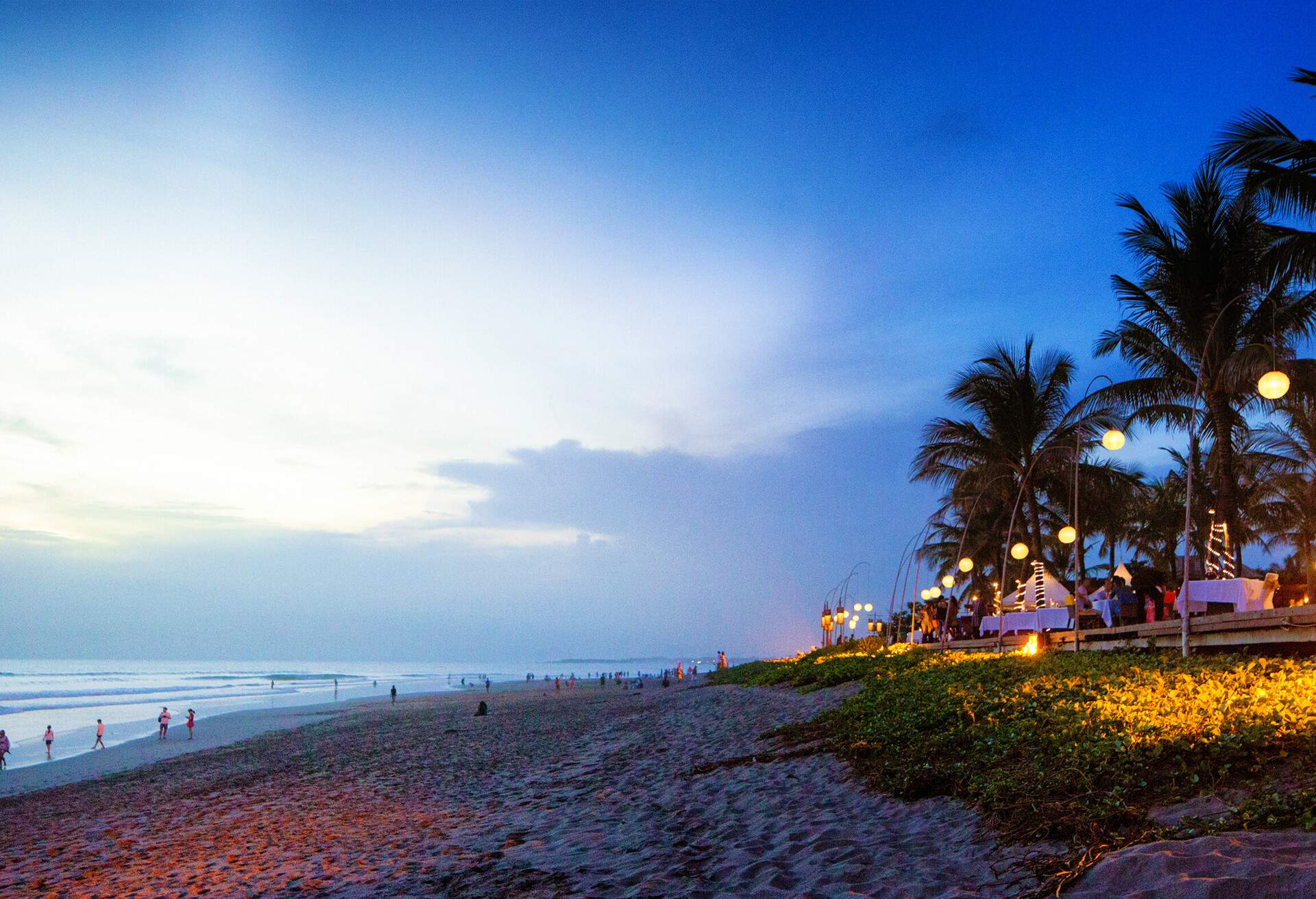 Seminyak Denpasar Bali Indonesia beach scene at sunset with a crowd at the left and a beach side restaurant at the right.