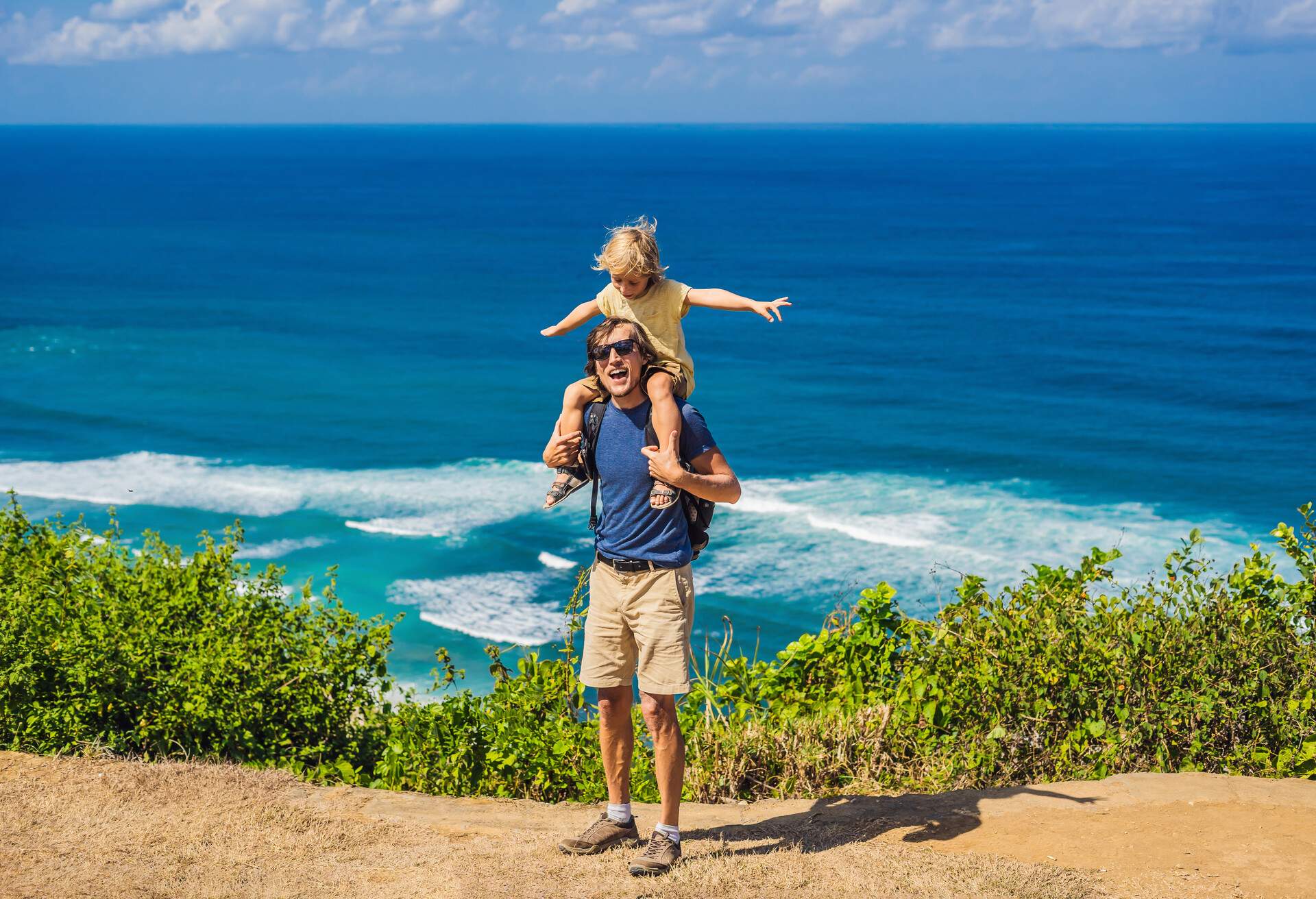 Dad and son travelers on a cliff above the beach. Empty paradise beach, blue sea waves in Bali island, Indonesia. Suluban and Nyang Nyang place. Traveling with kids concept.