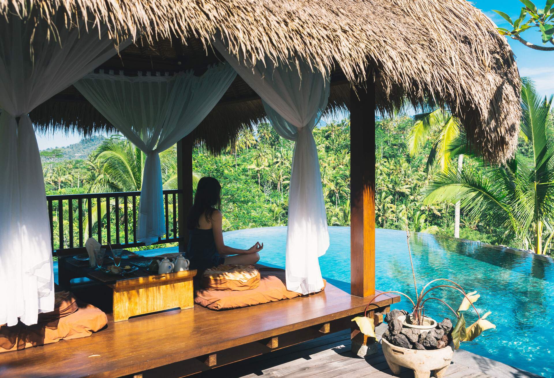 Girl meditating by the pool on a background of palms, Bali, Indonesia.The girl sits in a gazebo at a bali in a lotus pose and meditates with a view of the palms and the pool. Breakfast in Bali, Ubud.; Shutterstock ID 698319172; Purpose: Archives; Brand (KAYAK, Momondo, Any): Any