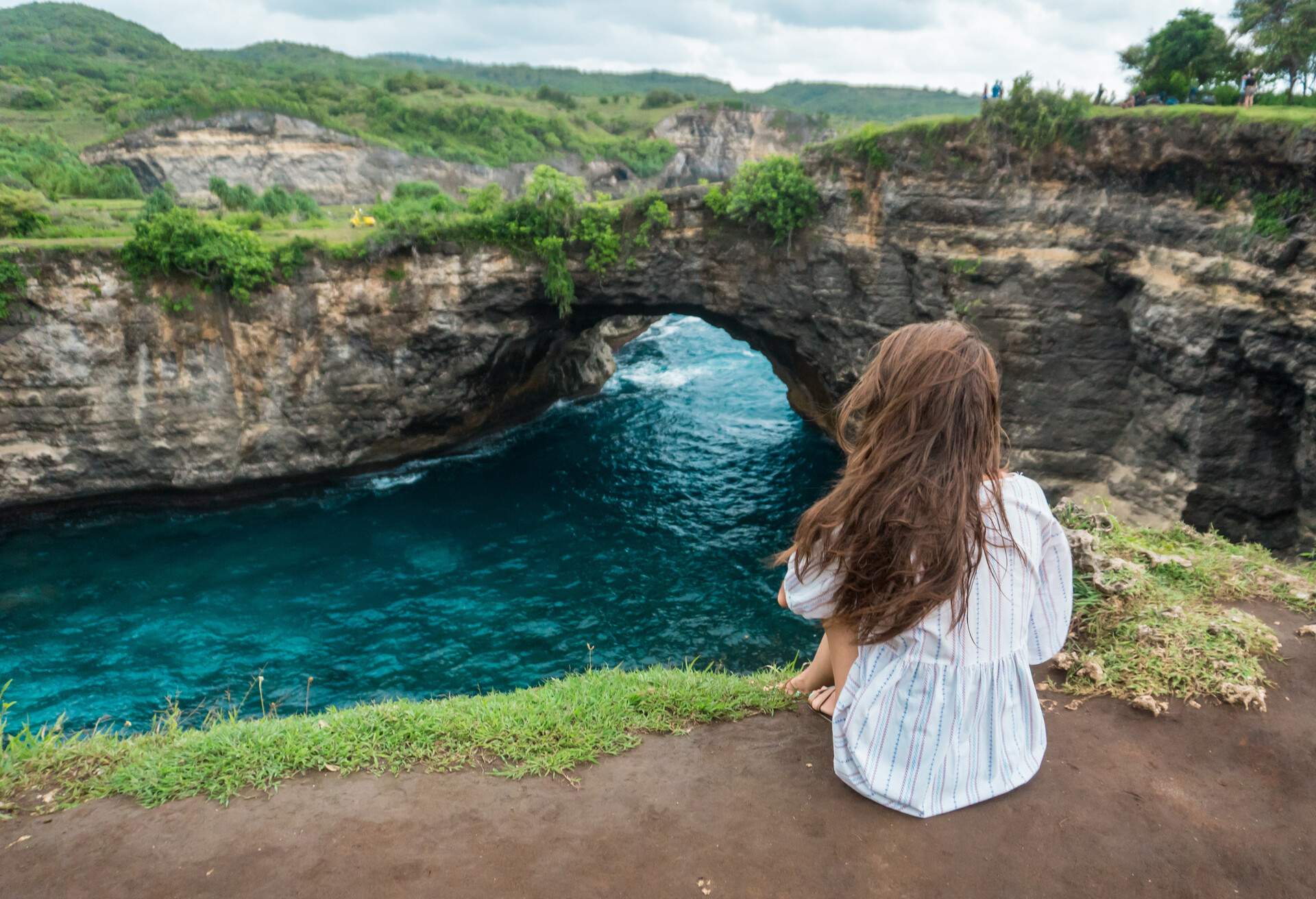 Woman sitting on cliff and enjoying beautiful landscape. Stone arch over the ocean. Broken beach, Nusa Penida, Indonesia