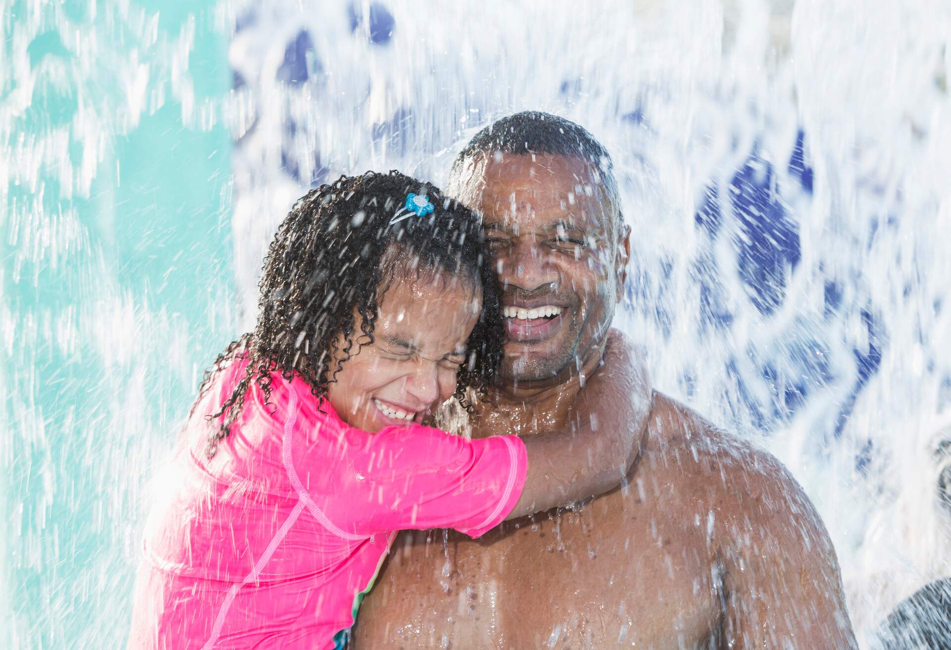 An African-American father having fun at the water park with his 7 year old mixed race daughter. The little girl's arms are wrapped around daddy's neck. They are under a waterfall smiling with their eyes shut tightly to keep the water out.