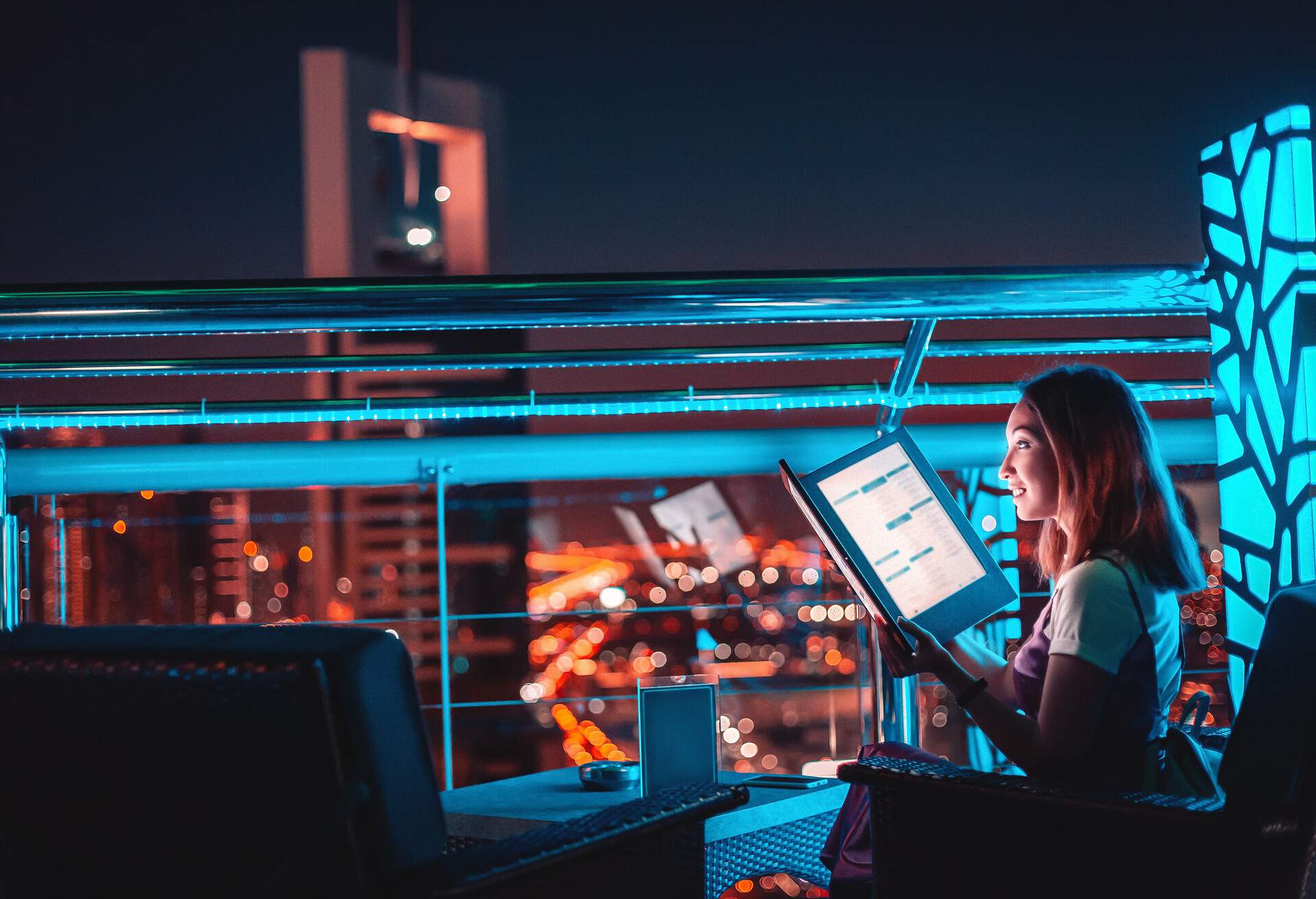 Asian girl reads the menu at the bar on the outdoor roof terrace overlooking the night city