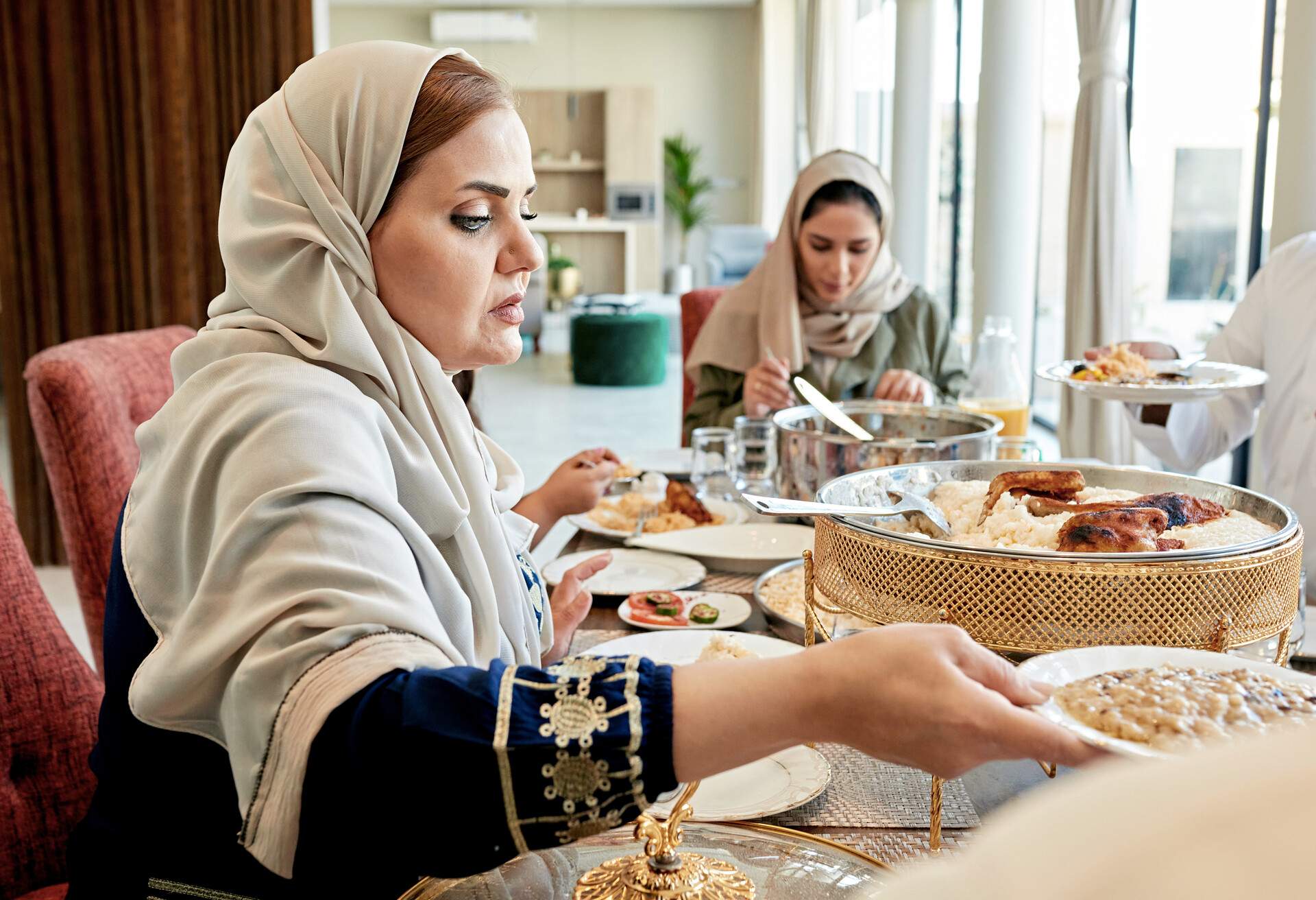 Side view of woman in abaya and hijab sitting at dining table in modern home with family members and passing plate.