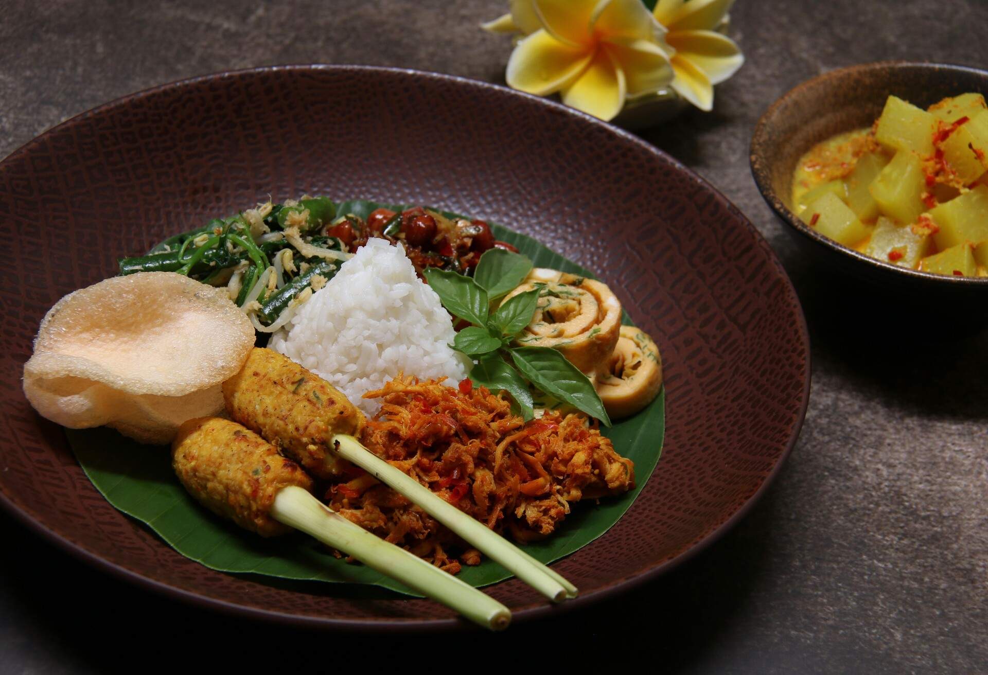 Nasi Campur Bali, the Balinese popular rice dish. Steamed white rice served with side dishes such as Sate Lilit (minced seafood satay), Ayam Pelalah (shredded chicken in red chili paste), Telur Dadar (egg omelet), Kacang Teri (peanuts and anchovies), Jukut Urap (vegetable salad with grated coconut dressing), Kerupuk Udang (prawn crackers) and Jukut Gedang Mekuah (green papaya curry) served separately in a bowl.