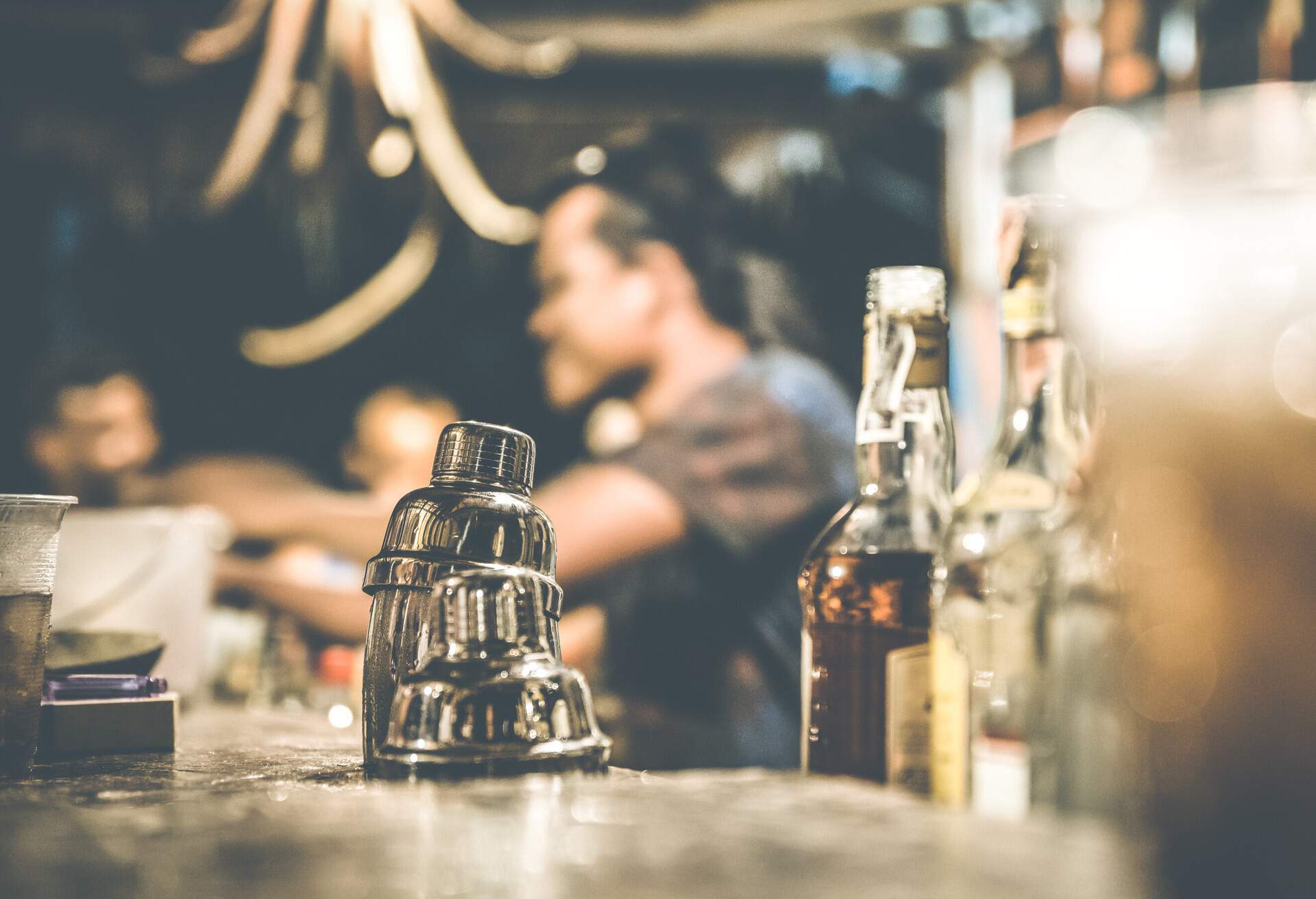 Blurred defocused side view of barman and guests drinking and having fun at cocktail bar - Social gathering concept with people enjoying time together - Warm retro contrast filter with focus on shaker; Shutterstock ID 586937294