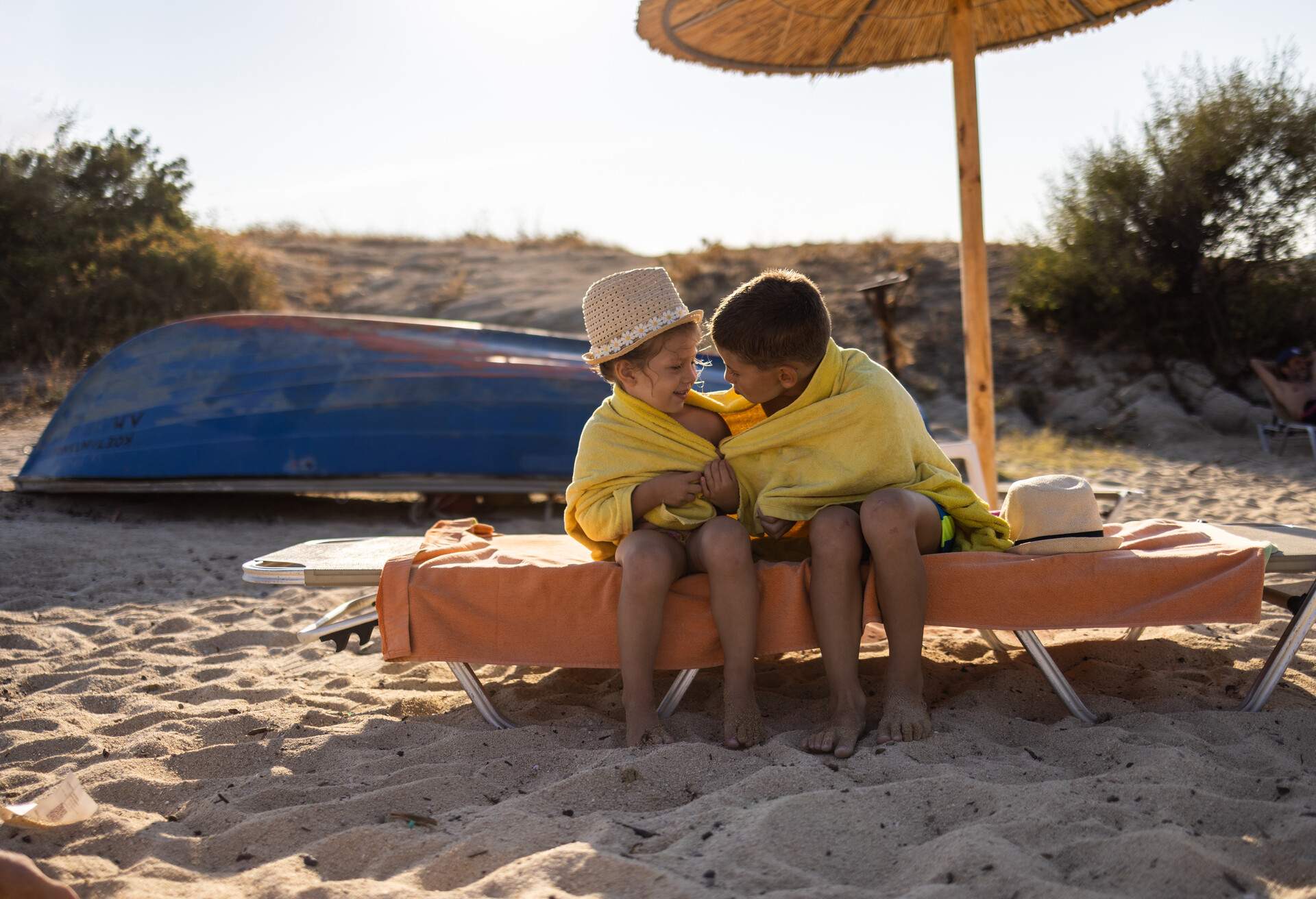 Little brother and sister wrapped in towels sitting on deck chair on beach together