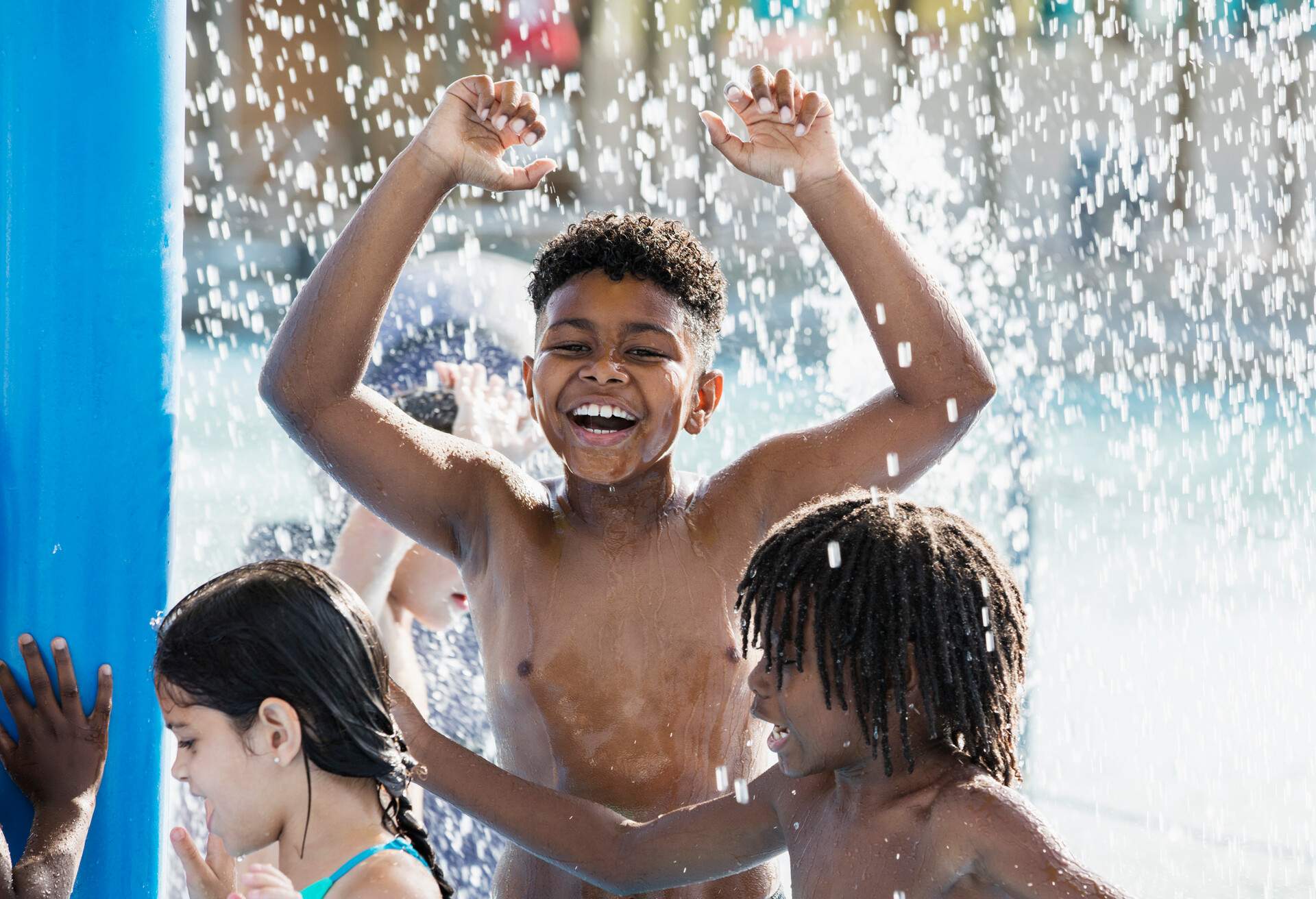 A 10 year old African-American boy having fun playing at a water park. He is standing under splashing water with a group of friends, arms raised, laughing and looking at the camera.