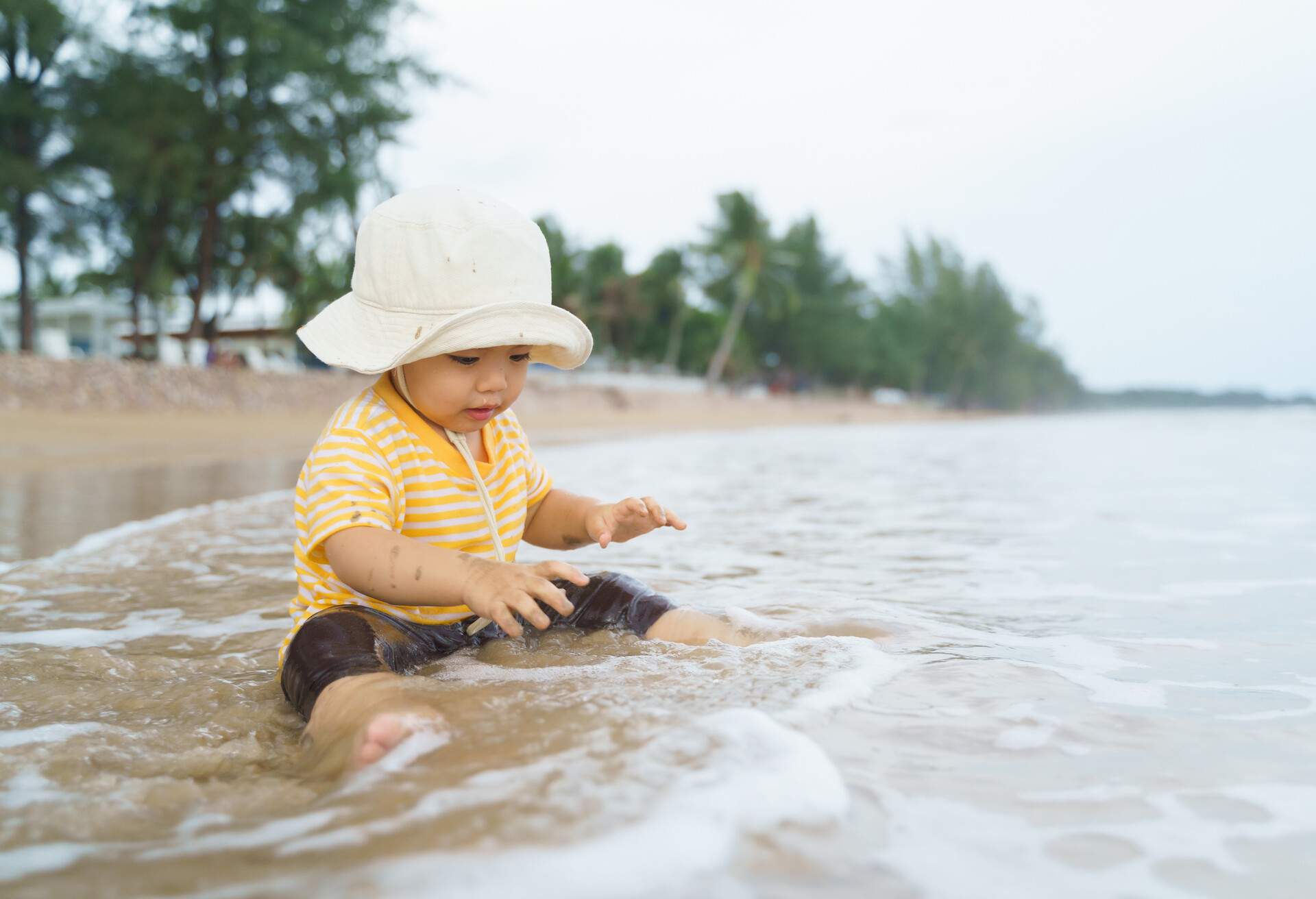 Asian adorable boy wearing hat sitting playing at the beach in the water