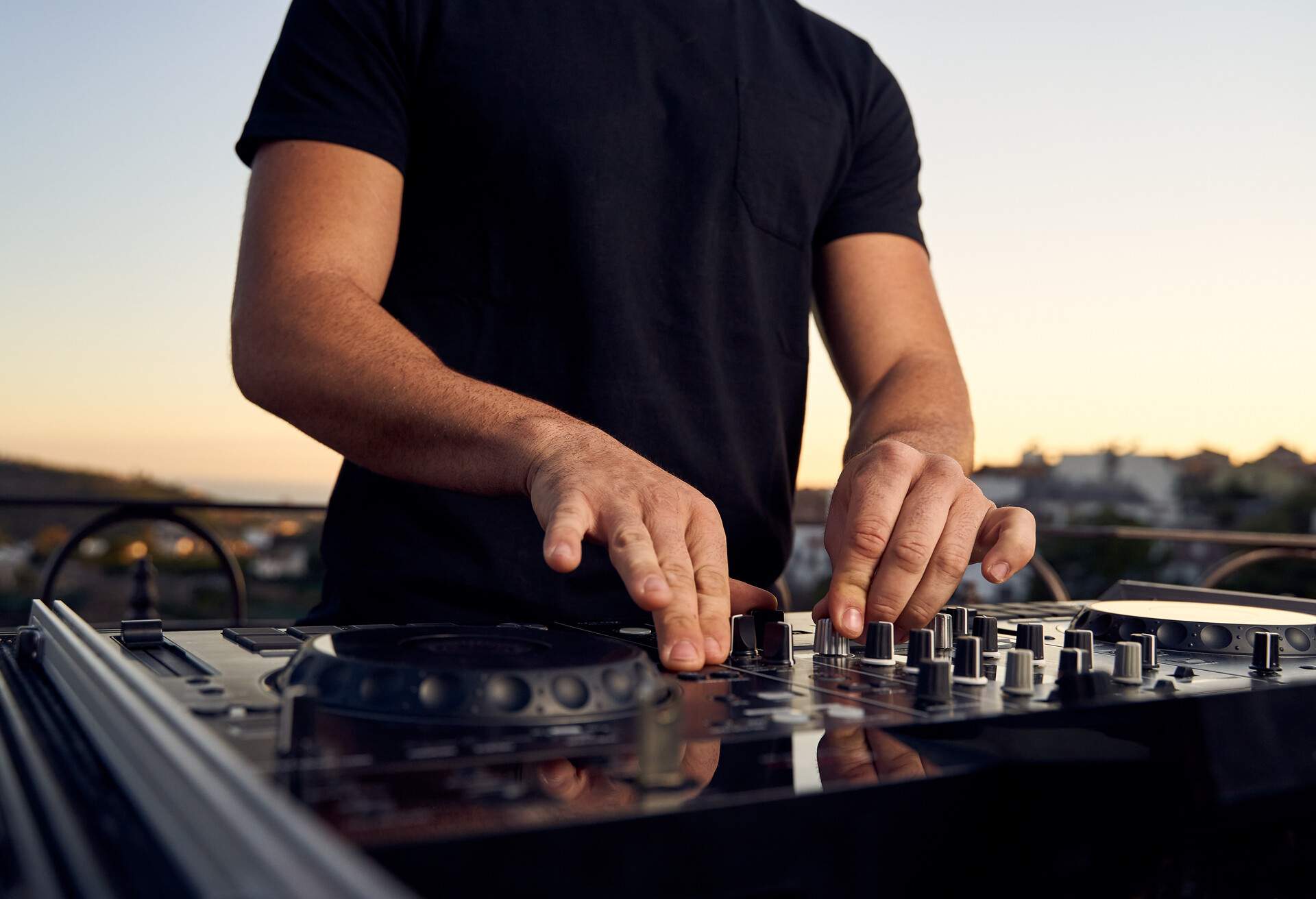 Unrecognizable male DJ hands playing music in a session with a mixer at a sunset party on a terrace.  Electronic music concept.