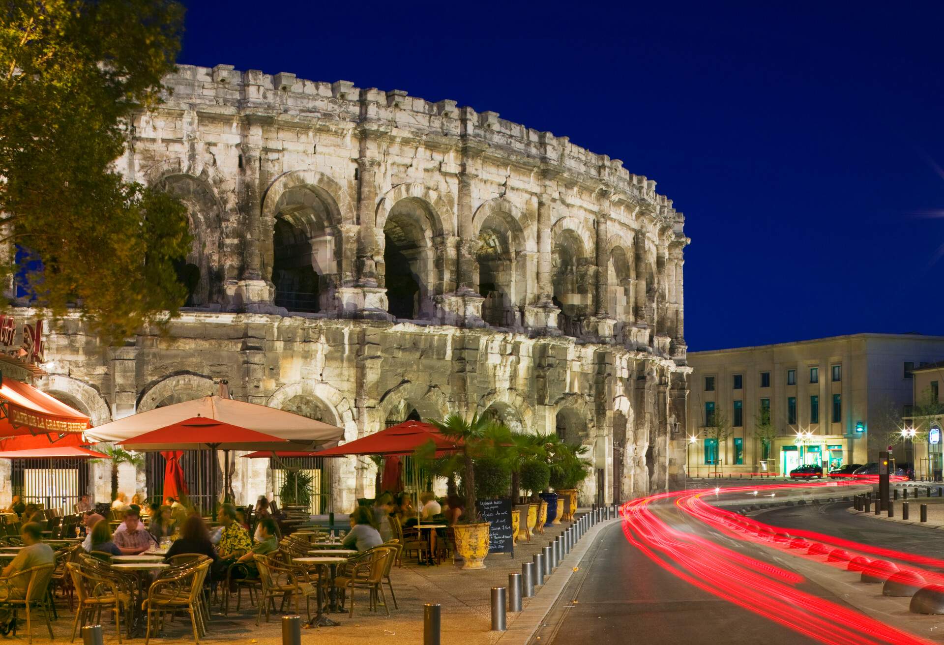 Cafe tables beneath Les Arenes, the Roman amphitheater, at dusk.