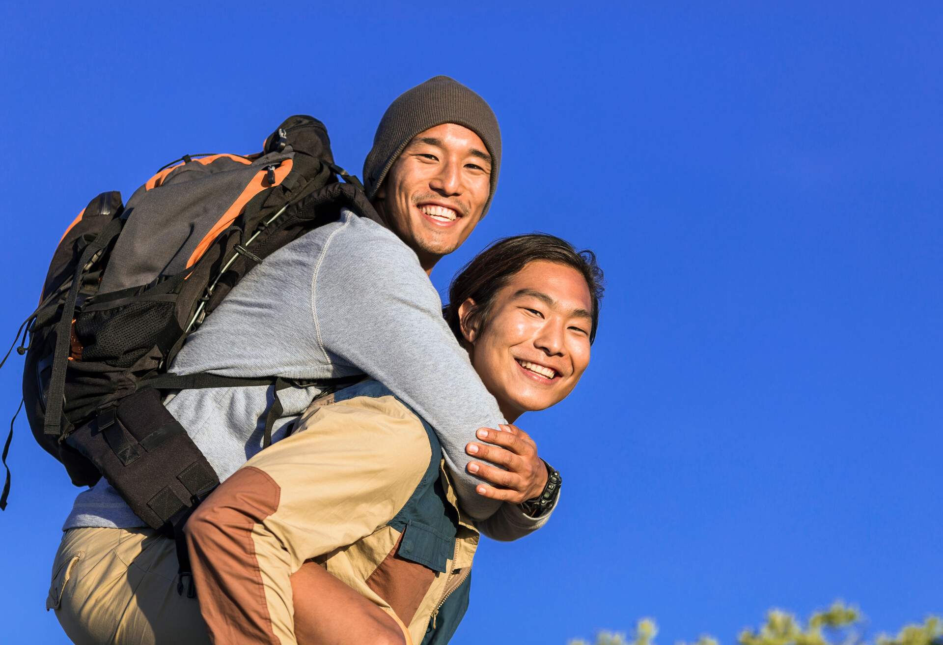 A photo of hiker giving piggyback ride to boyfriend. Portrait of happy men are against clear blue sky. Young males are in casuals. They are enjoying their weekends.