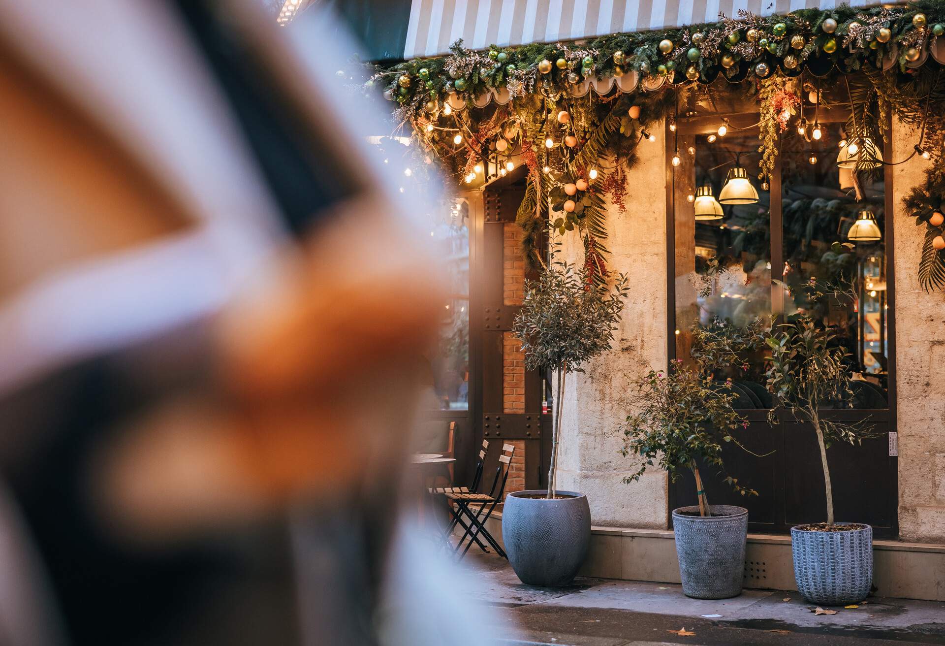 Close-up fancy Italian restaurant in the evening with Christmas decoration and lights at the top of the restaurant window, Pizza sign, plants standing in front of the Restaurant, Paris, woman with two baguettes in the forefront, focus on background, horizontal