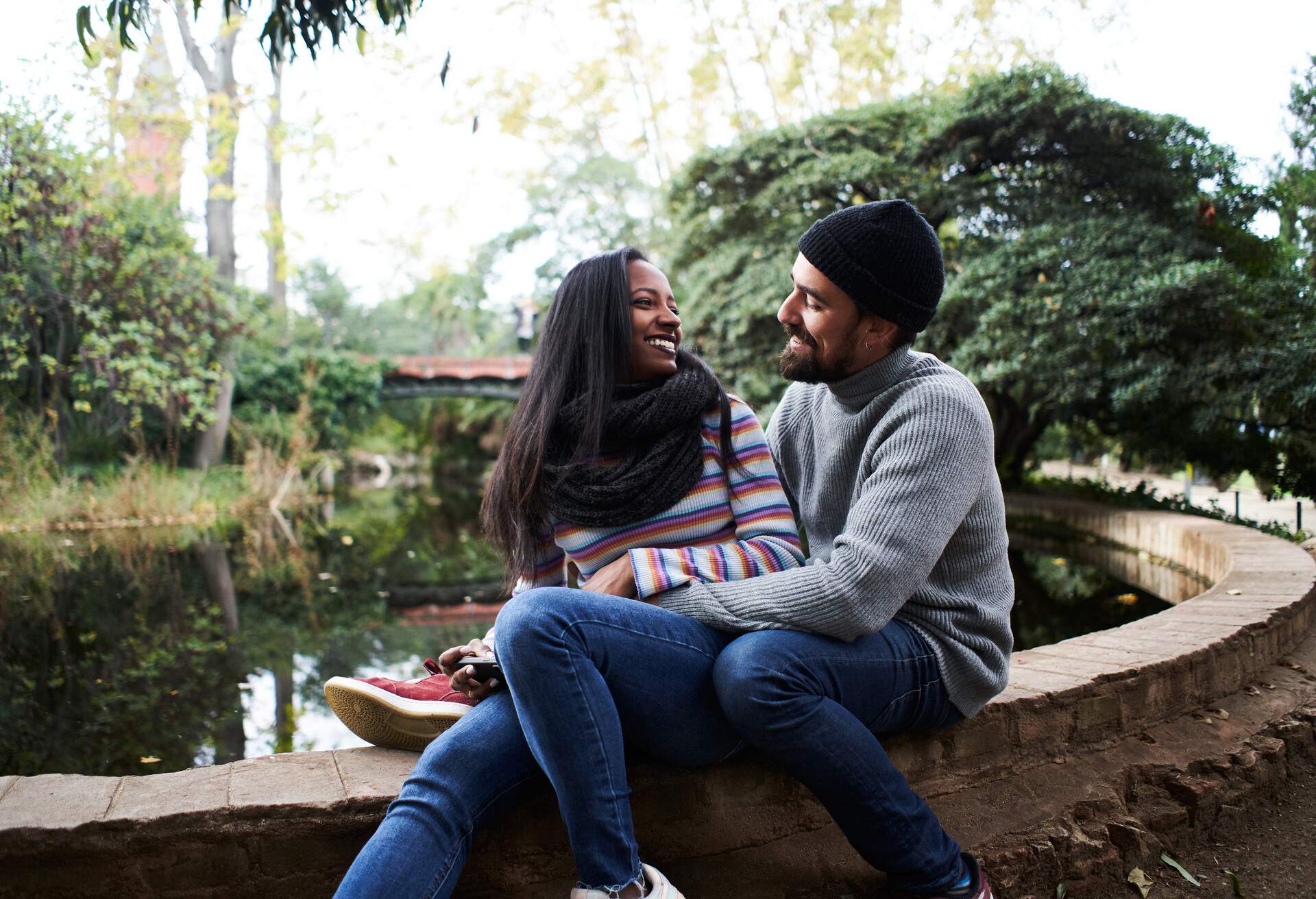 Mixed race couple sitting in a lake border while the male surround his female partner with his body and both look at each other in the eyes smiling.