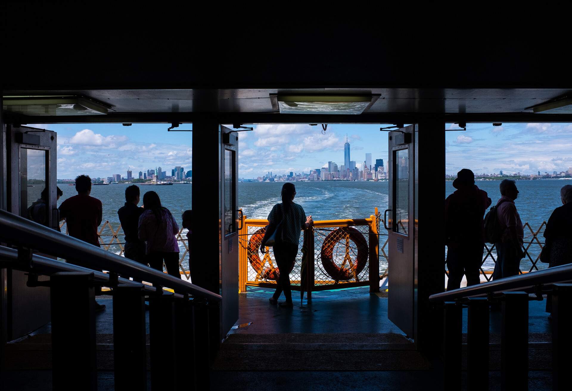 Silhouettes of passengers traveling from New York City aboard the Staten Island Ferry. The Manhattan skyline is seen through the windows in the distance.