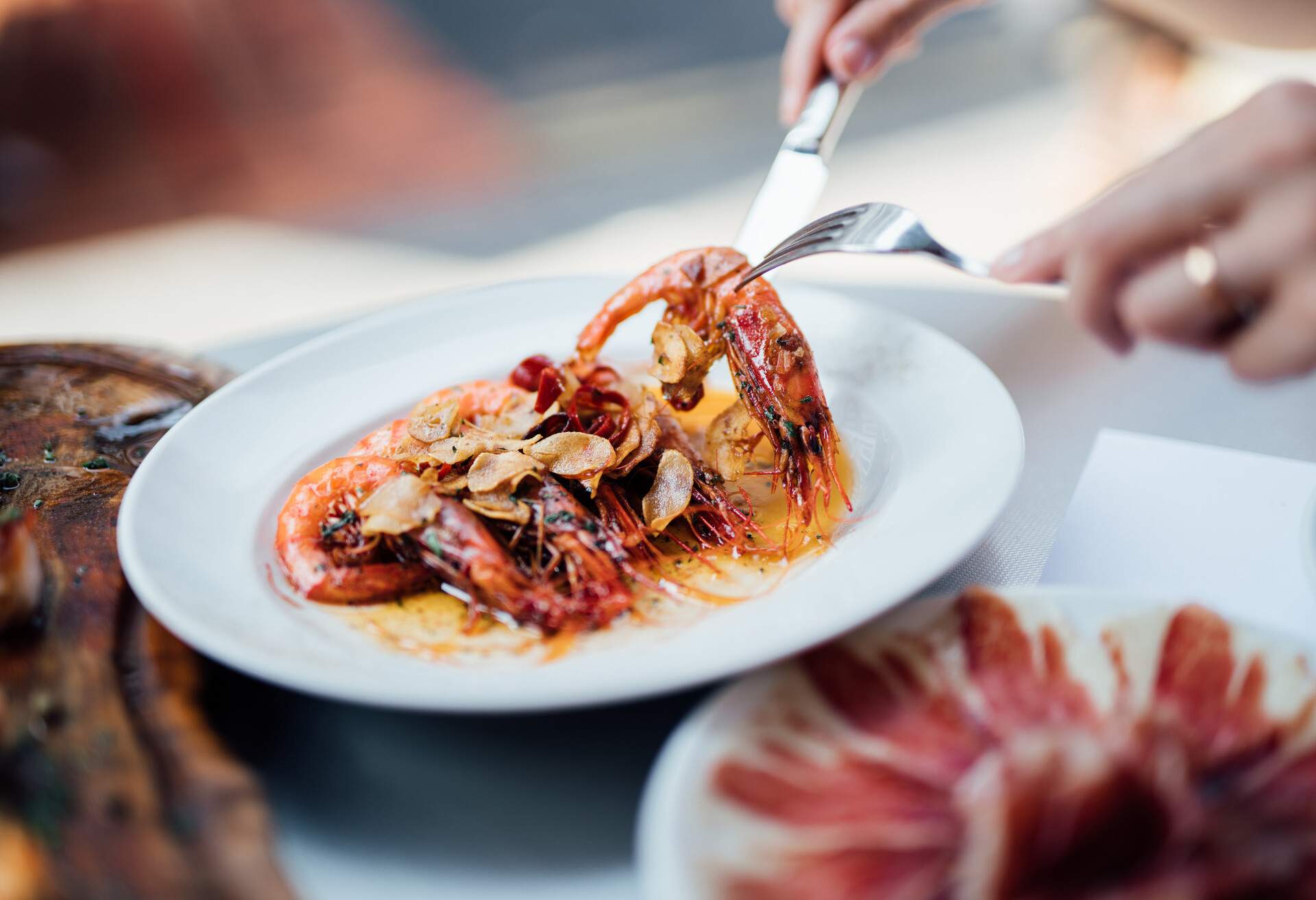 A plate of Spanish-style grilled shrimps, cooked with lemon juice, olive oil and sea salt. With Jamon Iberico in the background. Garlic prawn on a fork.