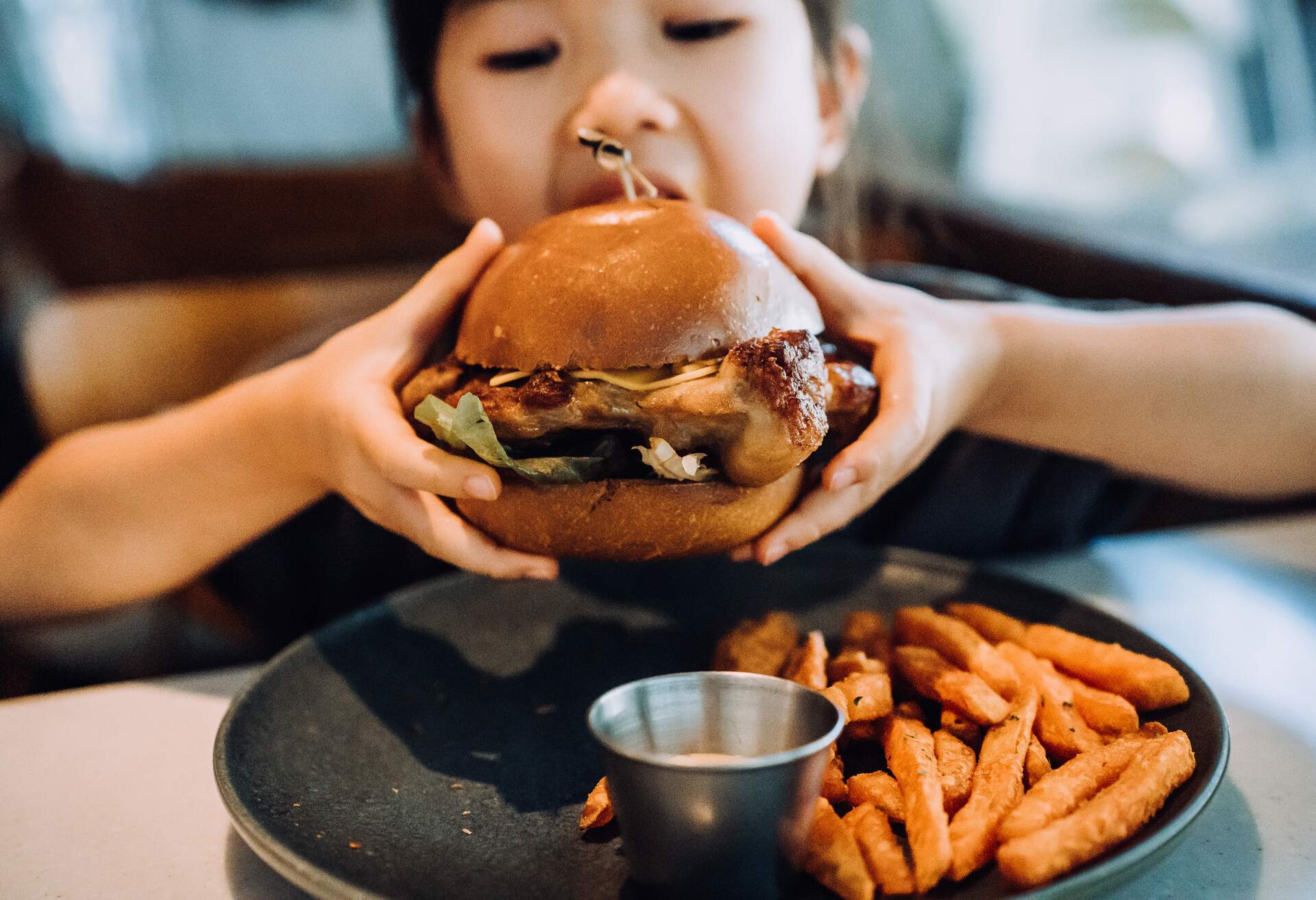 Close up shot of lovely little Asian girl enjoying eating a big burger with sweet potato fries in a restaurant. Eating out lifestyle