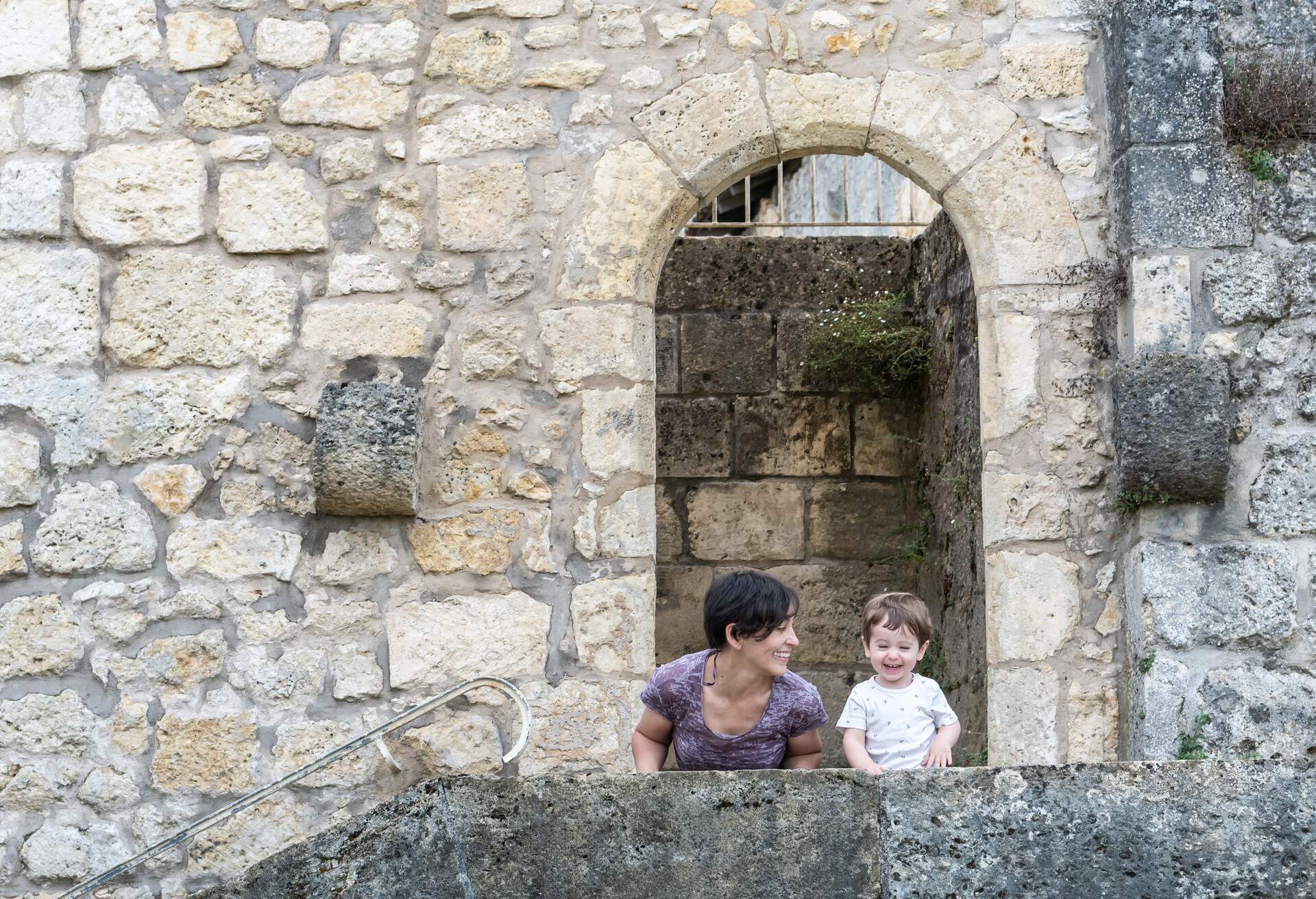 Mother looking at her little boy, both of them are smiling. They are crouched on a kerb of a staircase, this staircase takes you to the entrance of a park which you have to pass under a limestone arch. It belonged to an ancient castle and now belongs to the Sainte Marie church in the village of Roquefort.