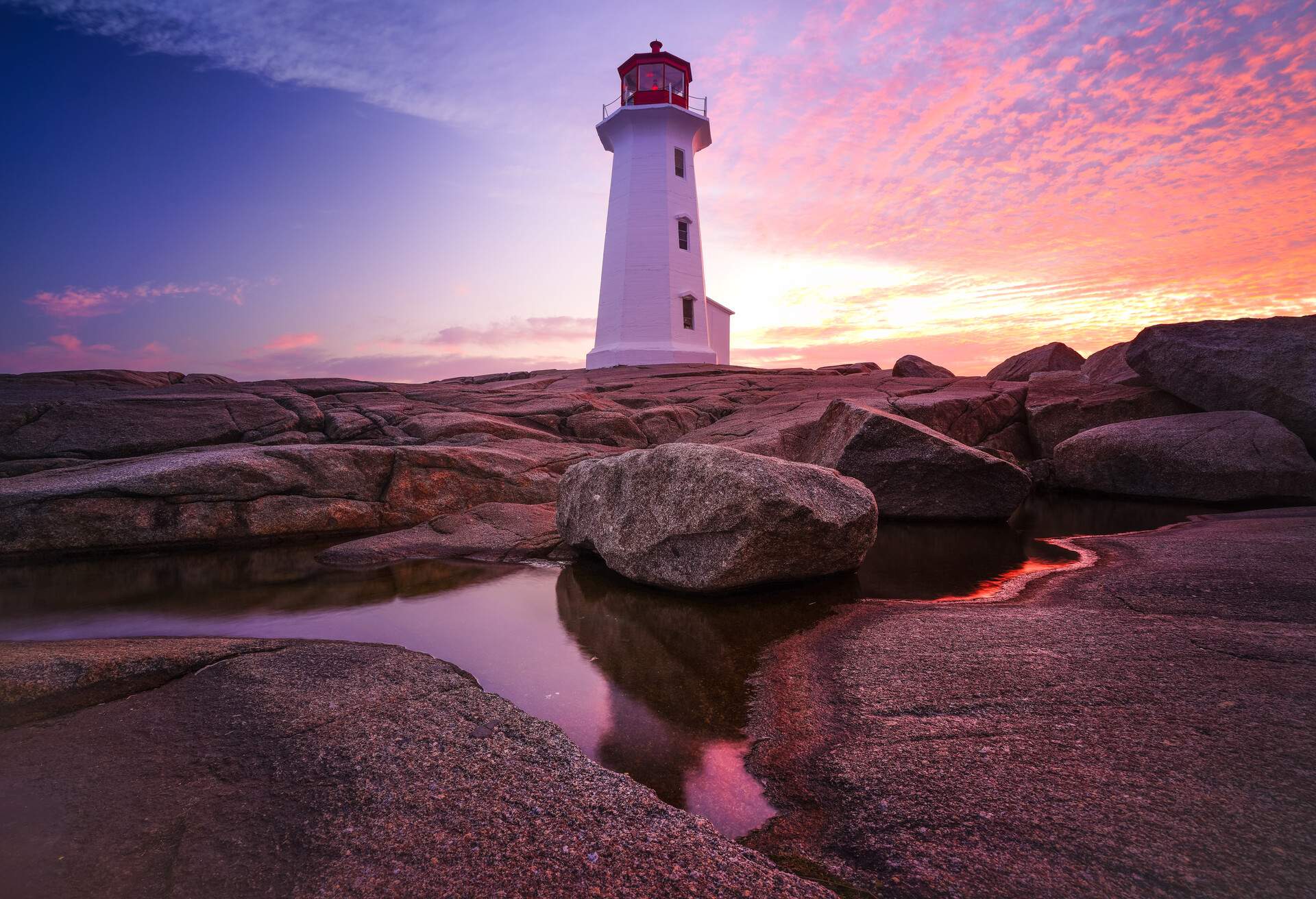 Peggys Point Lighthouse during a pink and purple sunrise, with mackerel clouds, reflected in a pool of water on the granite rock. Peggy's Cove, Nova Scotia.