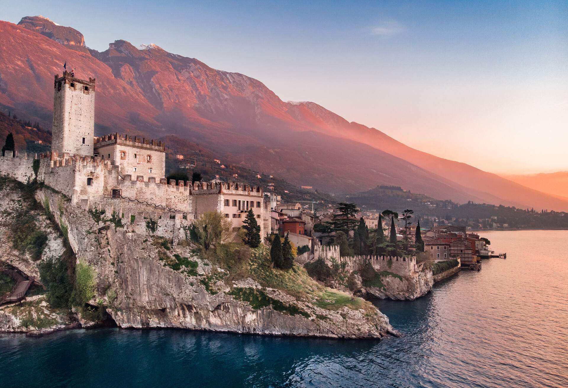 A medieval town with a castle above the rocky coast of a lake with a view of mountains in the background.