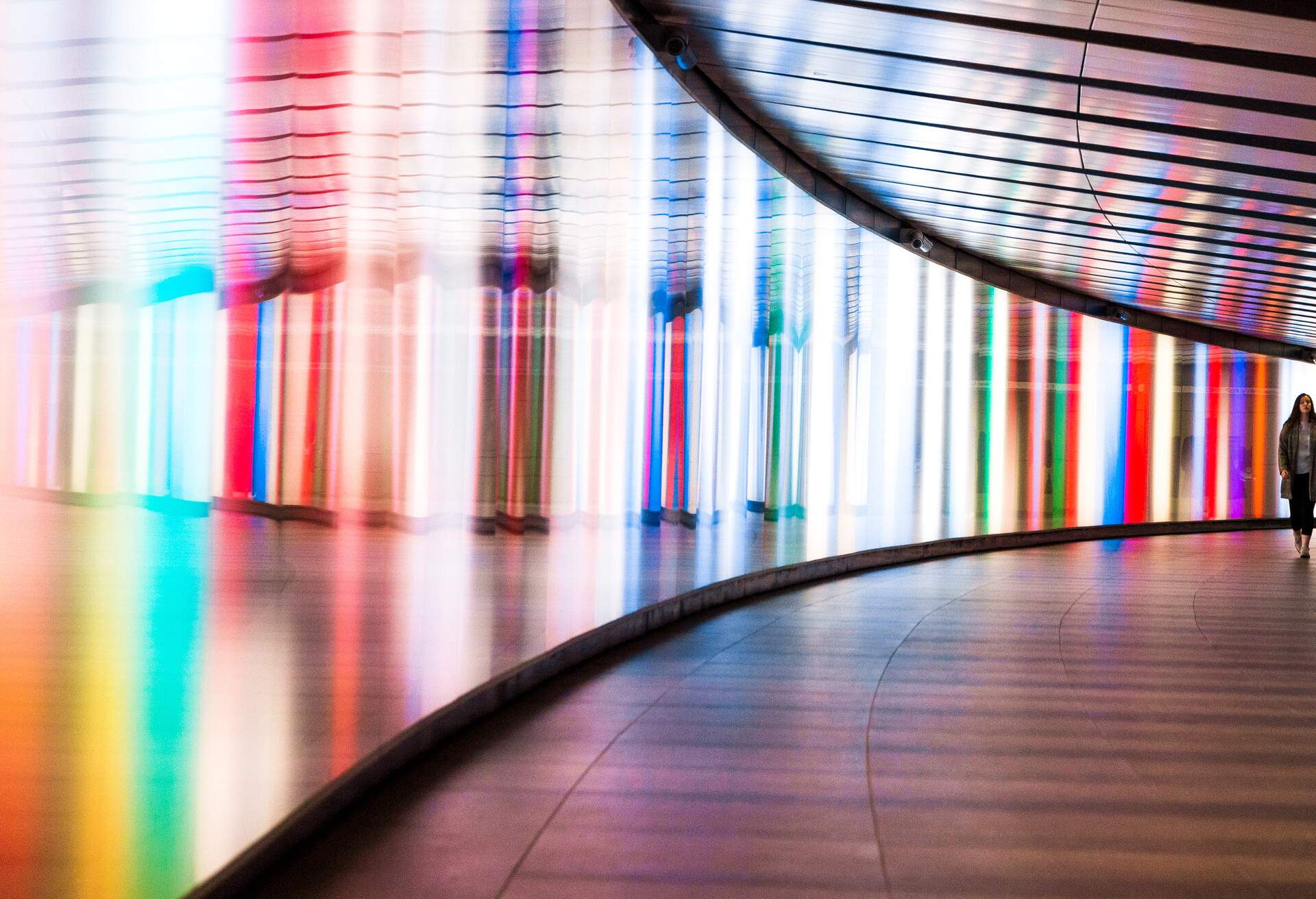 Wide angle color image depicting the front view of a young woman walking through a futuristic subway tunnel in central London, UK. The tunnel is illuminated with multi colored lights. The woman is in the far distance, and the focus is on the cool science fiction architecture of the subway tunnel that curves round into the distance. Room for copy space.
