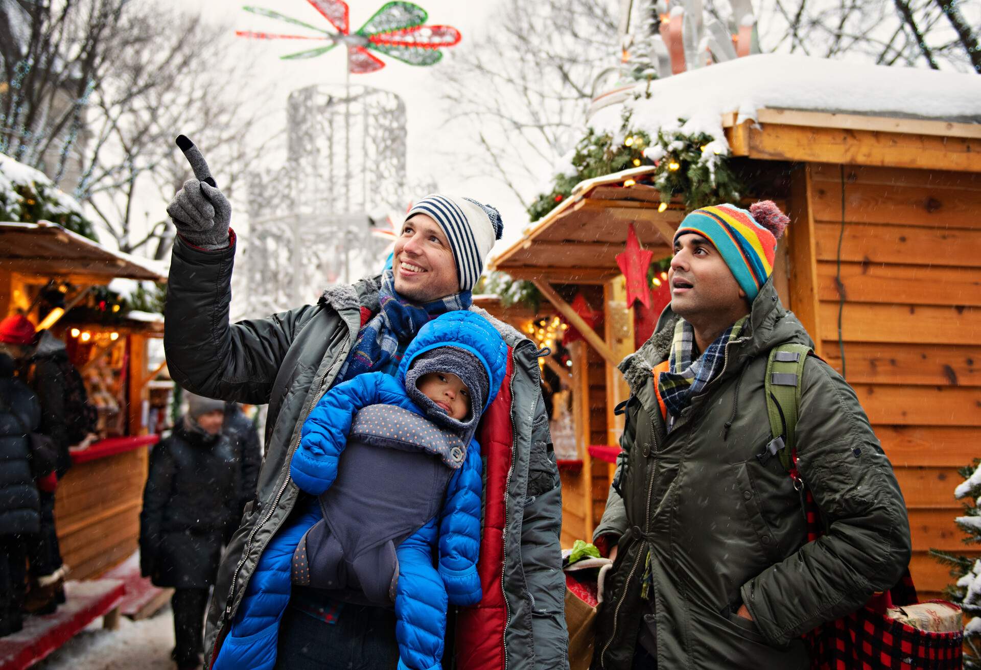 A couple with their child on a baby carrier backpack, wearing their winter clothes, on a snowy Christmas market.
