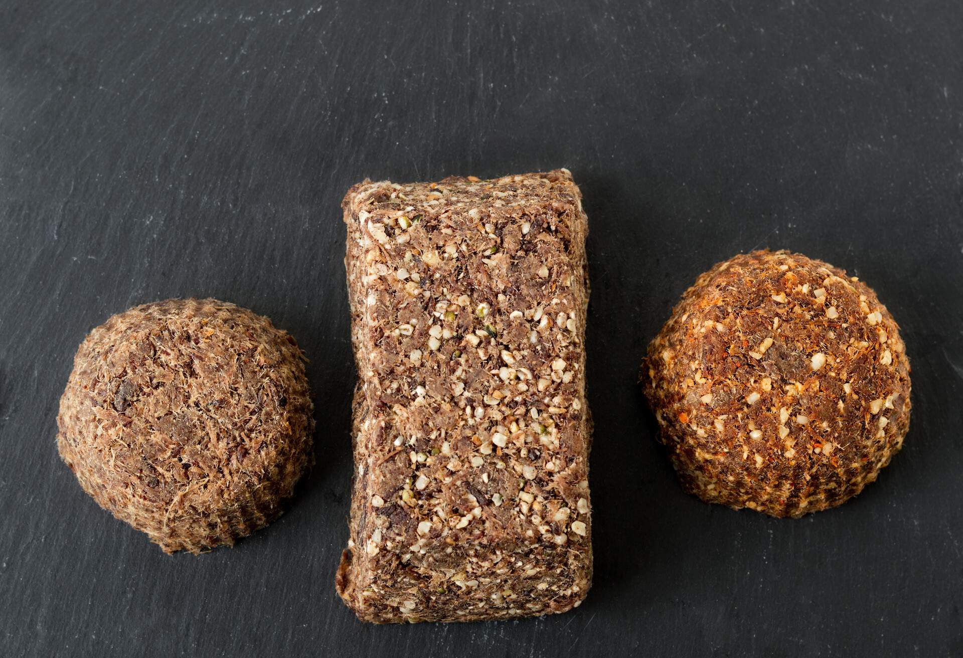 Top view of three bars of homemade beef meat and liver pemmican, plain with salt, with hemp seeds and hemp seeds and spices on a gray slate.