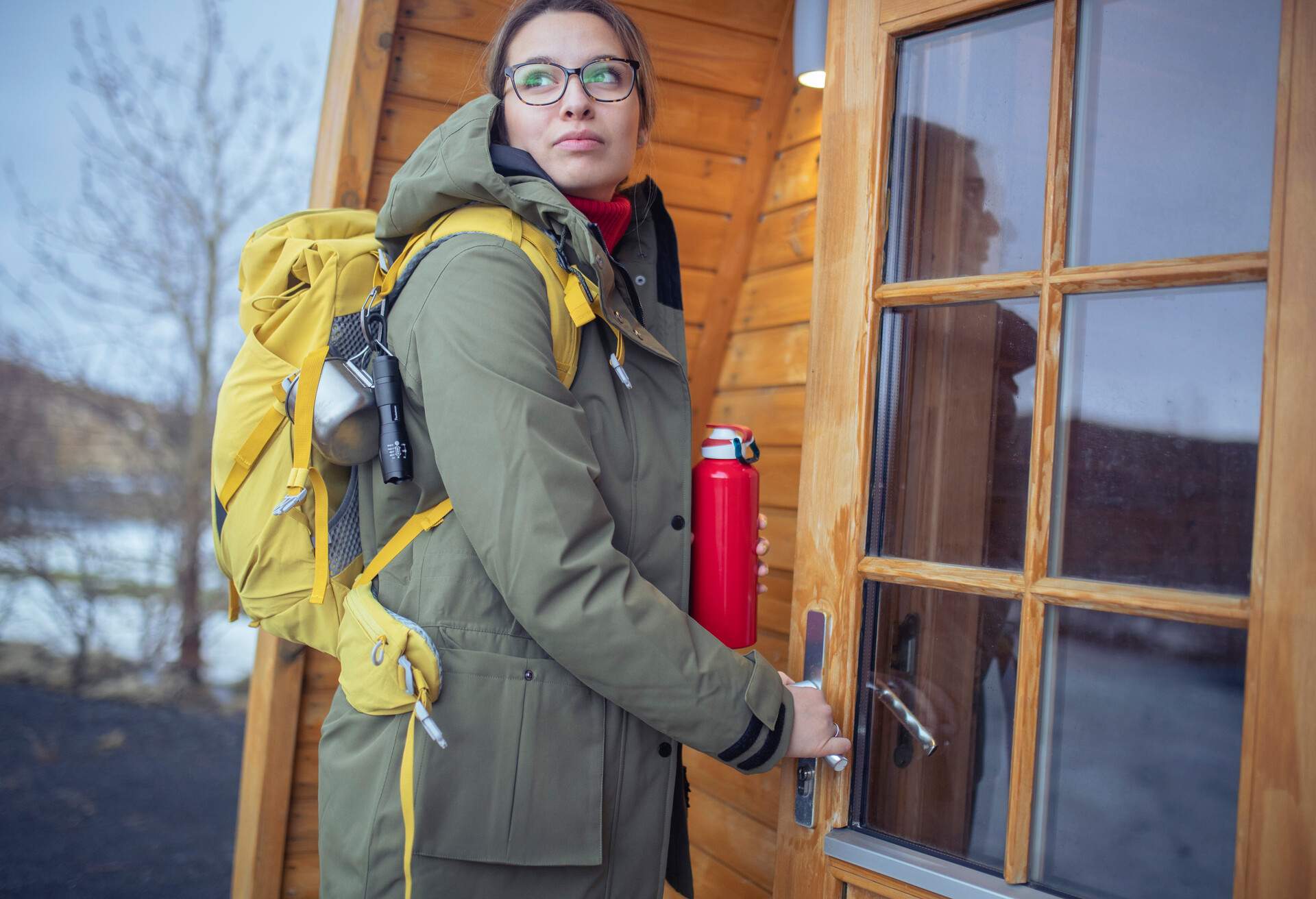 Woman holding a thermal flask about to open the door to get inside the wooden cabin.