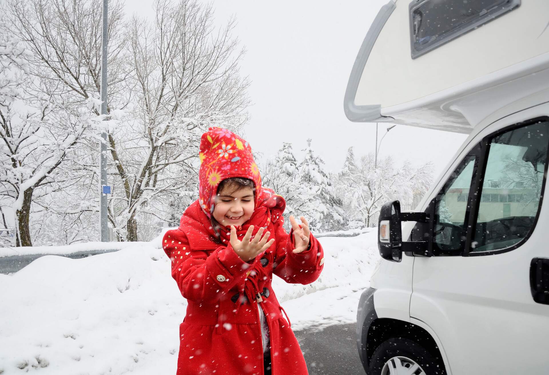 A young girl in a red coat and winter hat catching falling snowflakes next to a white campervan.