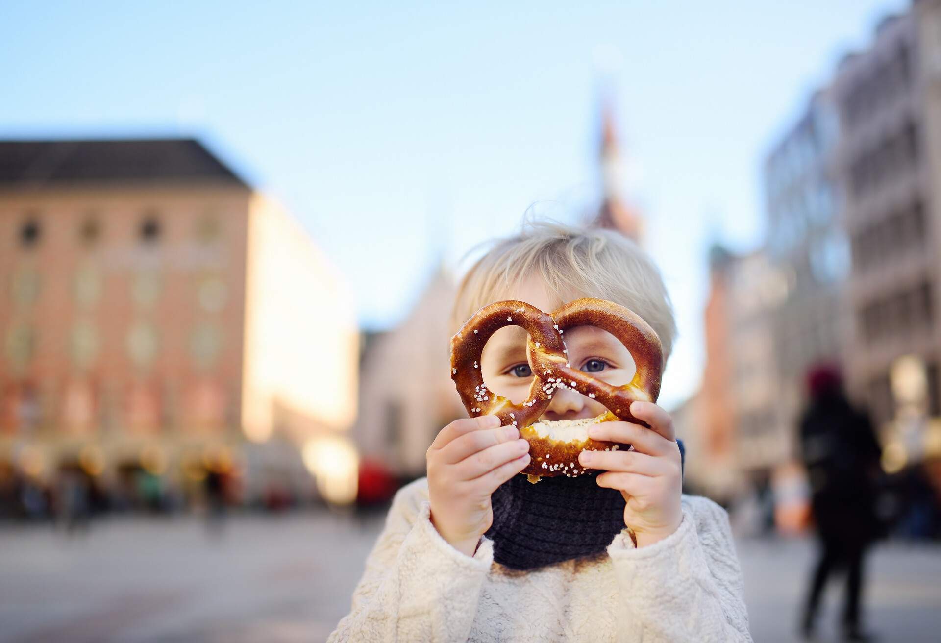 Little tourist holding traditional bavarian bread called pretzel on the town hall building background in Munich, Germany. Preschooler boy enjoy travel with his parents