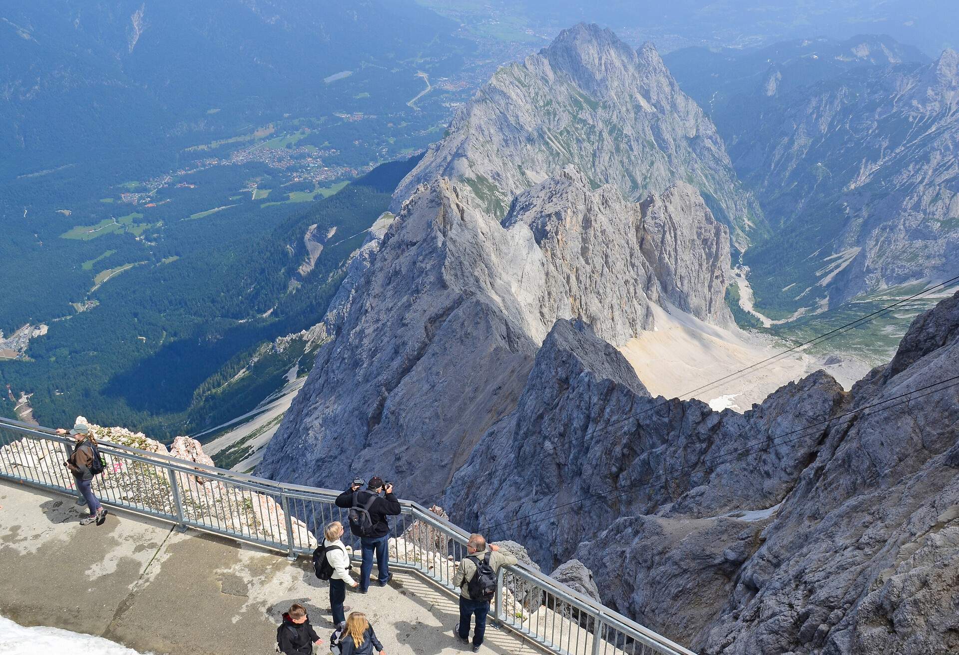 People walking and taking photos beside the side railings of a cliff overlooking a rocky mountain range and its nearby meadows.