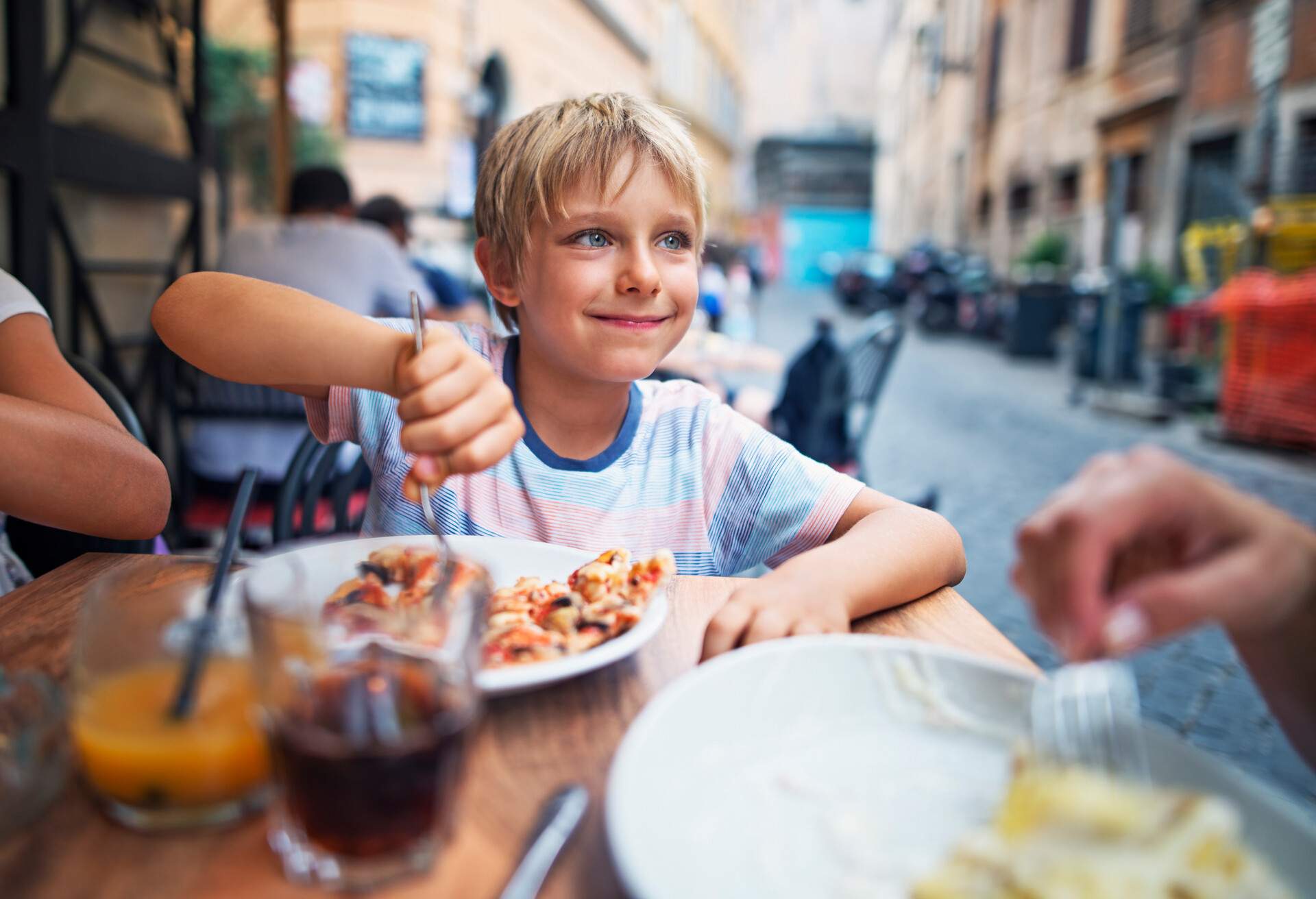 Little boy aged 7 having lunch with family in street restaurant in Rome, Italy. The girl is smiling and and looking sideways while getting a piece of pizza on the fork. Family is enjoying a happy meal together.