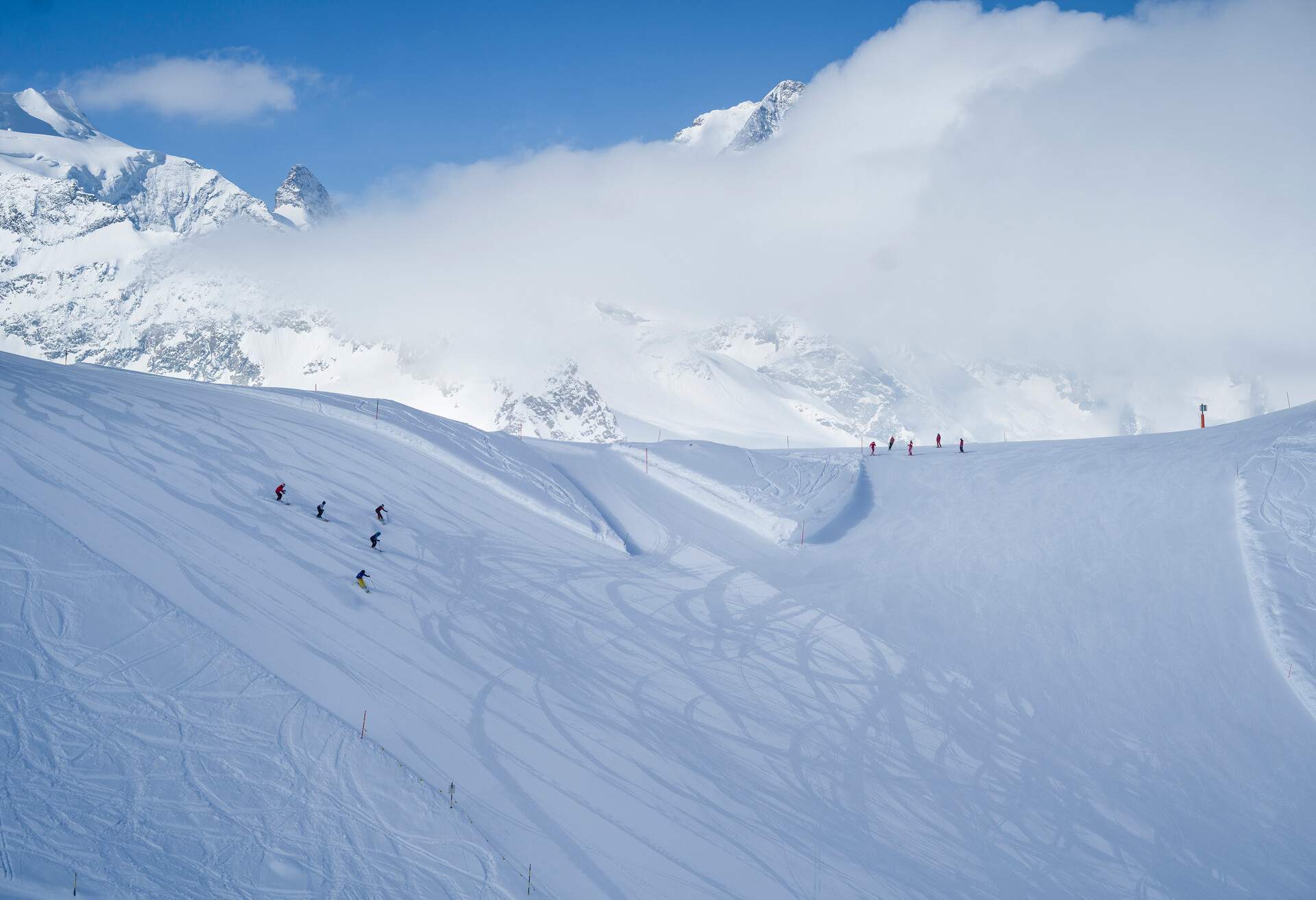 Skiers descend on a piste with a view of mountains covered in snow and a sea of clouds.