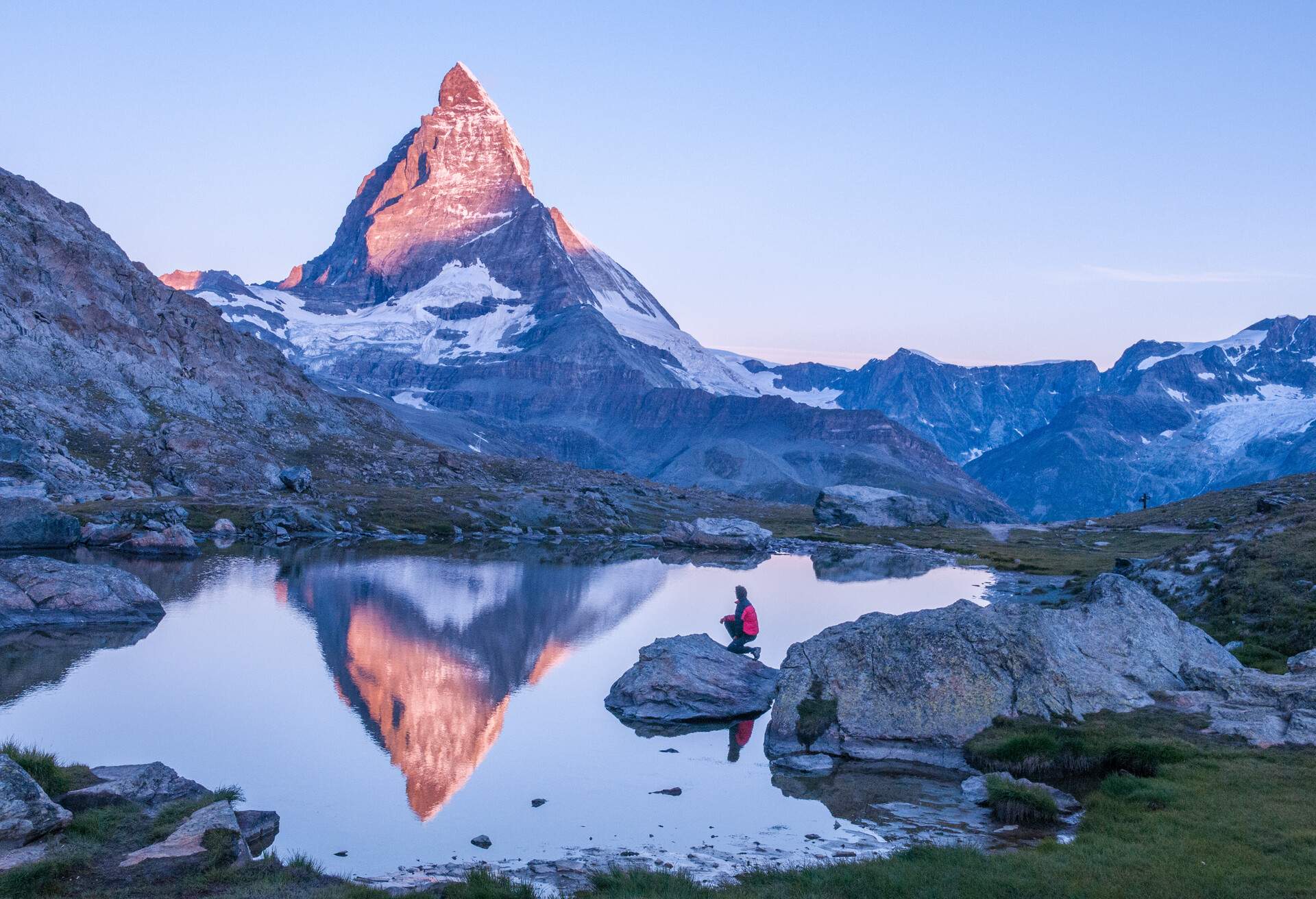 Early morning dawn scene of sunrise on the Matterhorn Mountain reflecting pink in the lake with male man on rock with red down jacket with clear blue sky Gornergrat Zermatt Matterhorn Europe