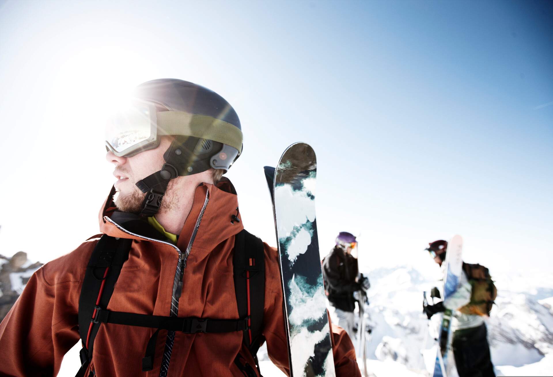 A group of skiers standing on a ski area, one in the foreground looking sideways, and two chatting in the background.