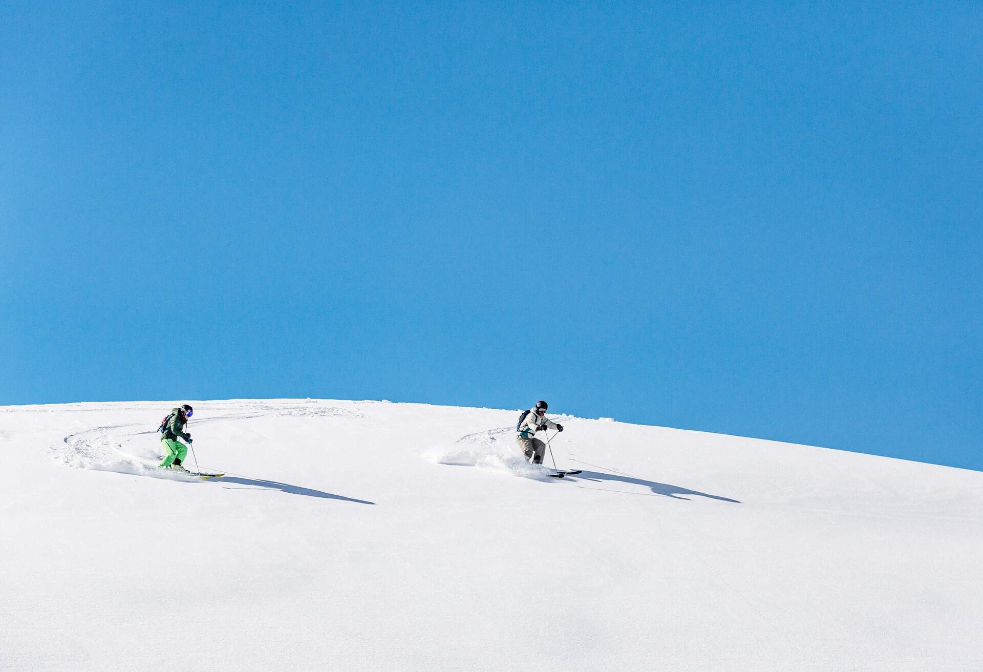 Two people skiing down an off-piste slope covered in fresh snow under the blue sky.