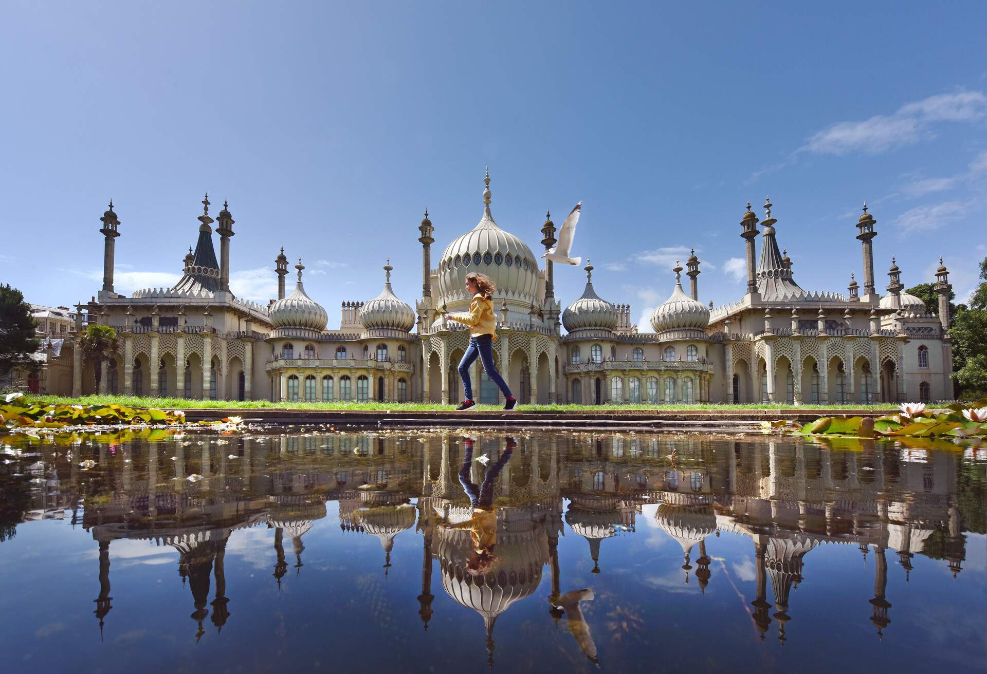A teenage girl reflected from across a pond - runs pursued by a seagull past the seaside town's landmark Georgian Royal Pavilion with it's famous domes and minarets