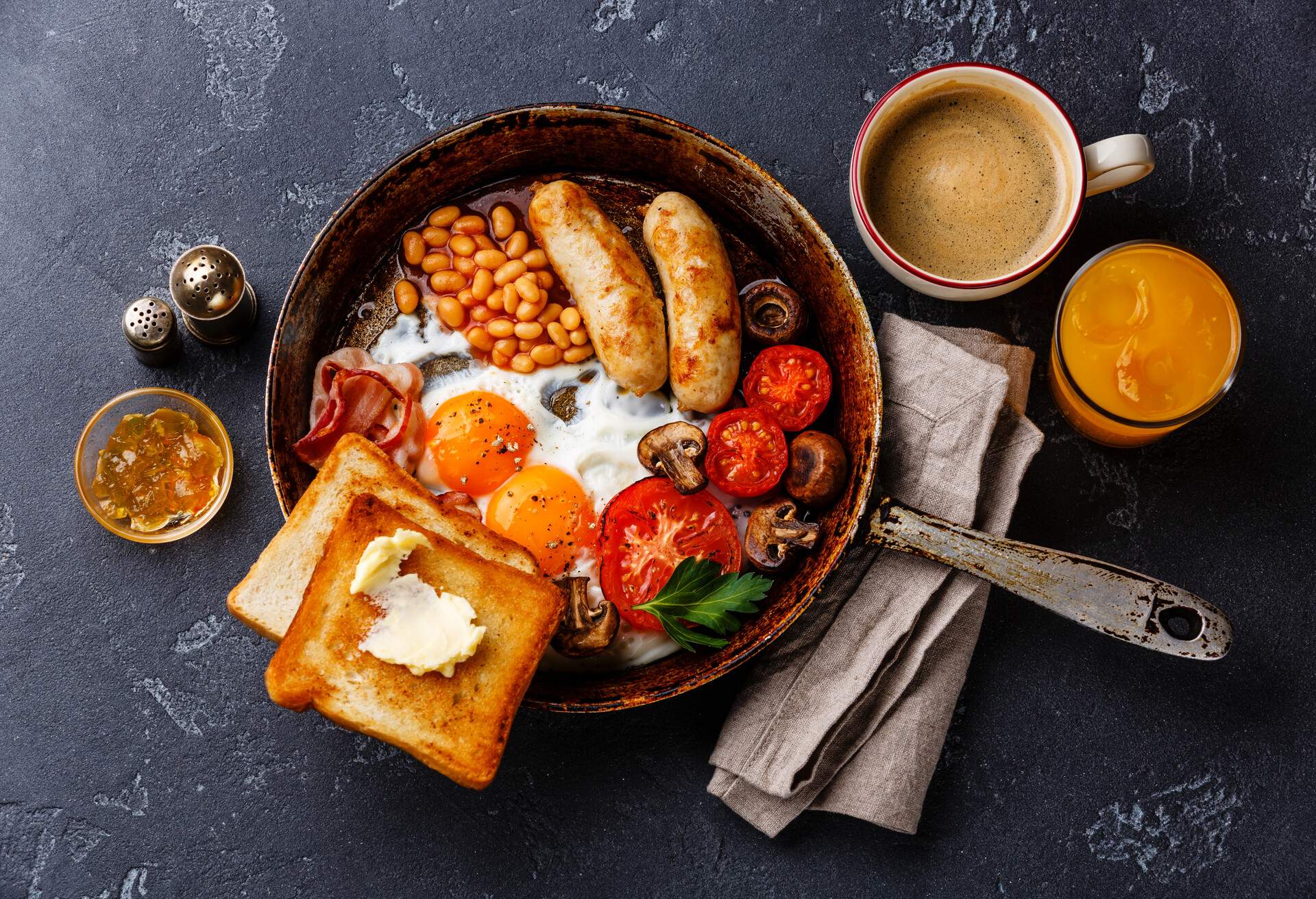 English breakfast in pan with fried eggs, sausages, bacon, beans, toasts and coffee on dark stone background