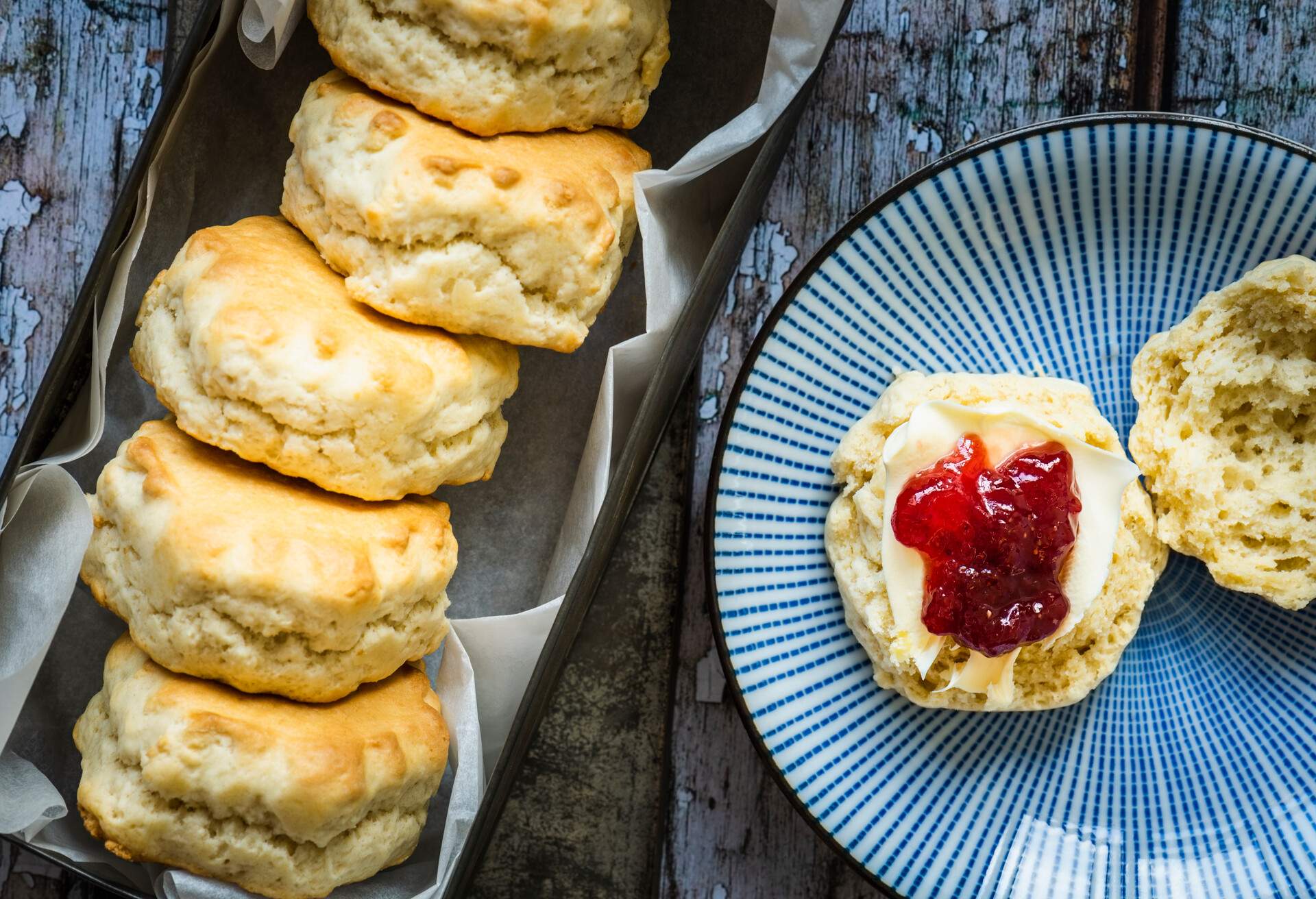 A freshly baked scone on a blue plate smothered in clotted cream and raspberry jam. 