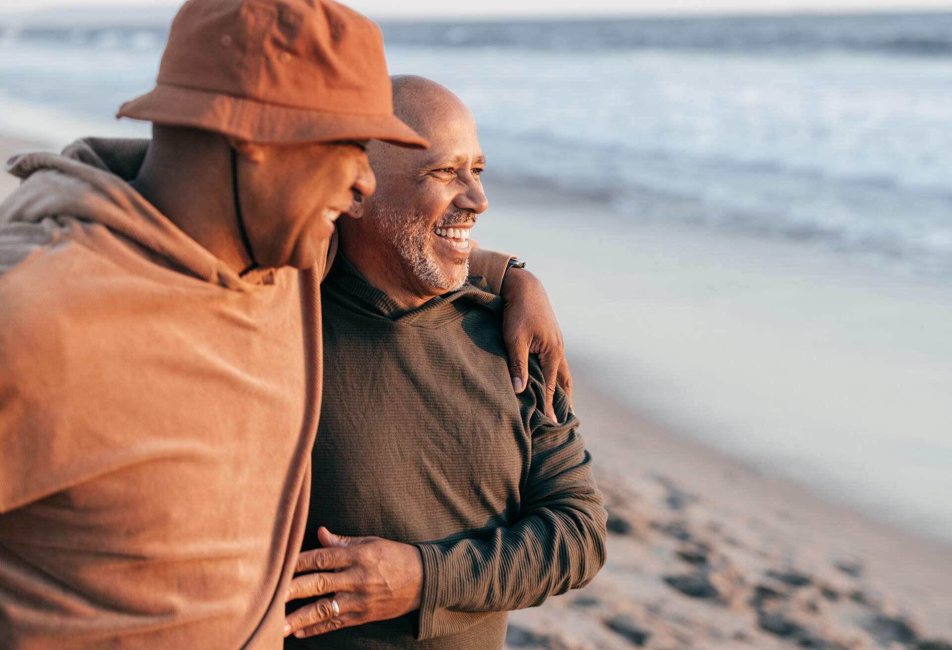 Elderly male lovers smile as they wrap their arms over each other's shoulders on the beach.