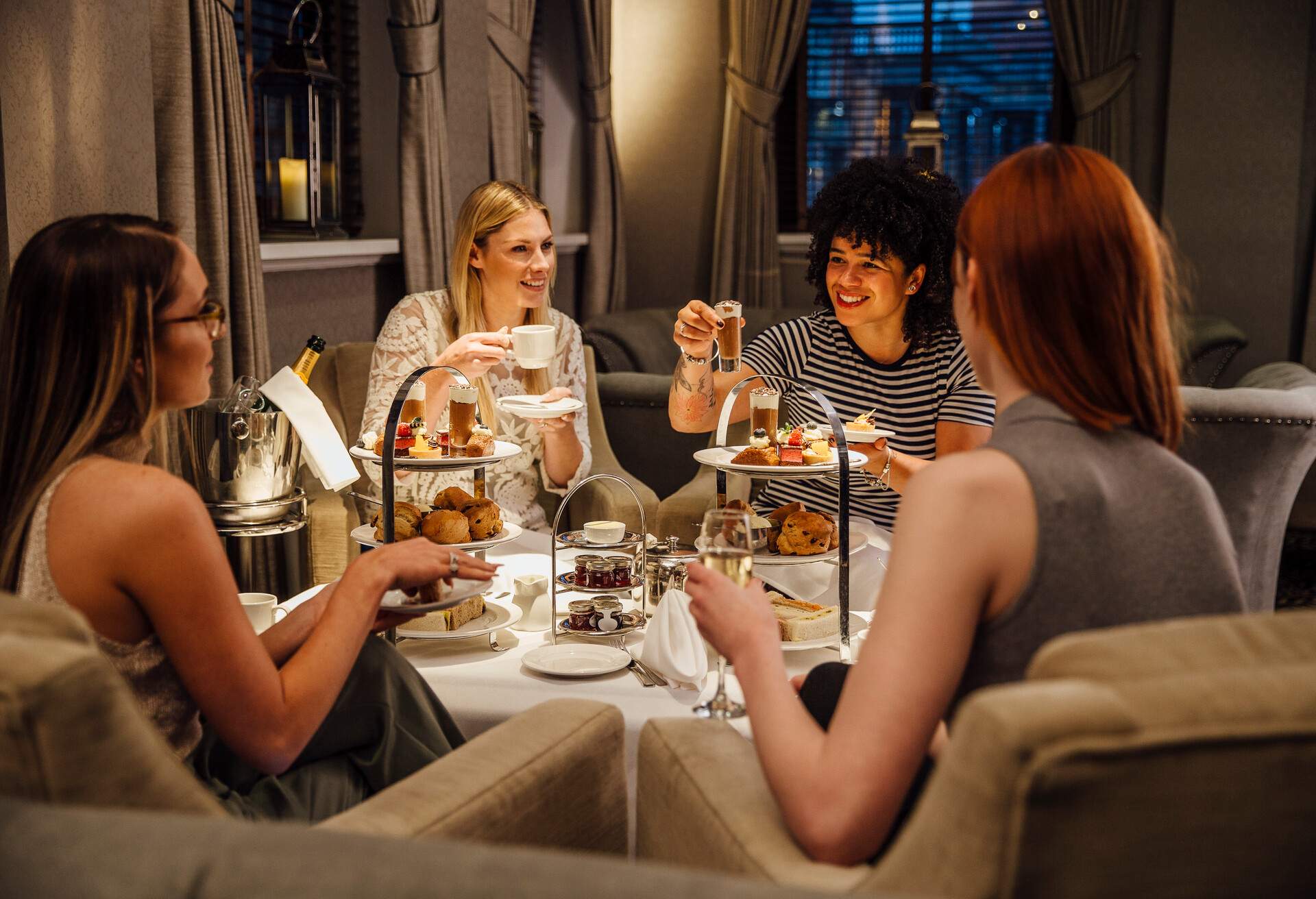 Four women are sitting together enjoying afternoon tea. They are talking and eating and they have champagne and tea. 